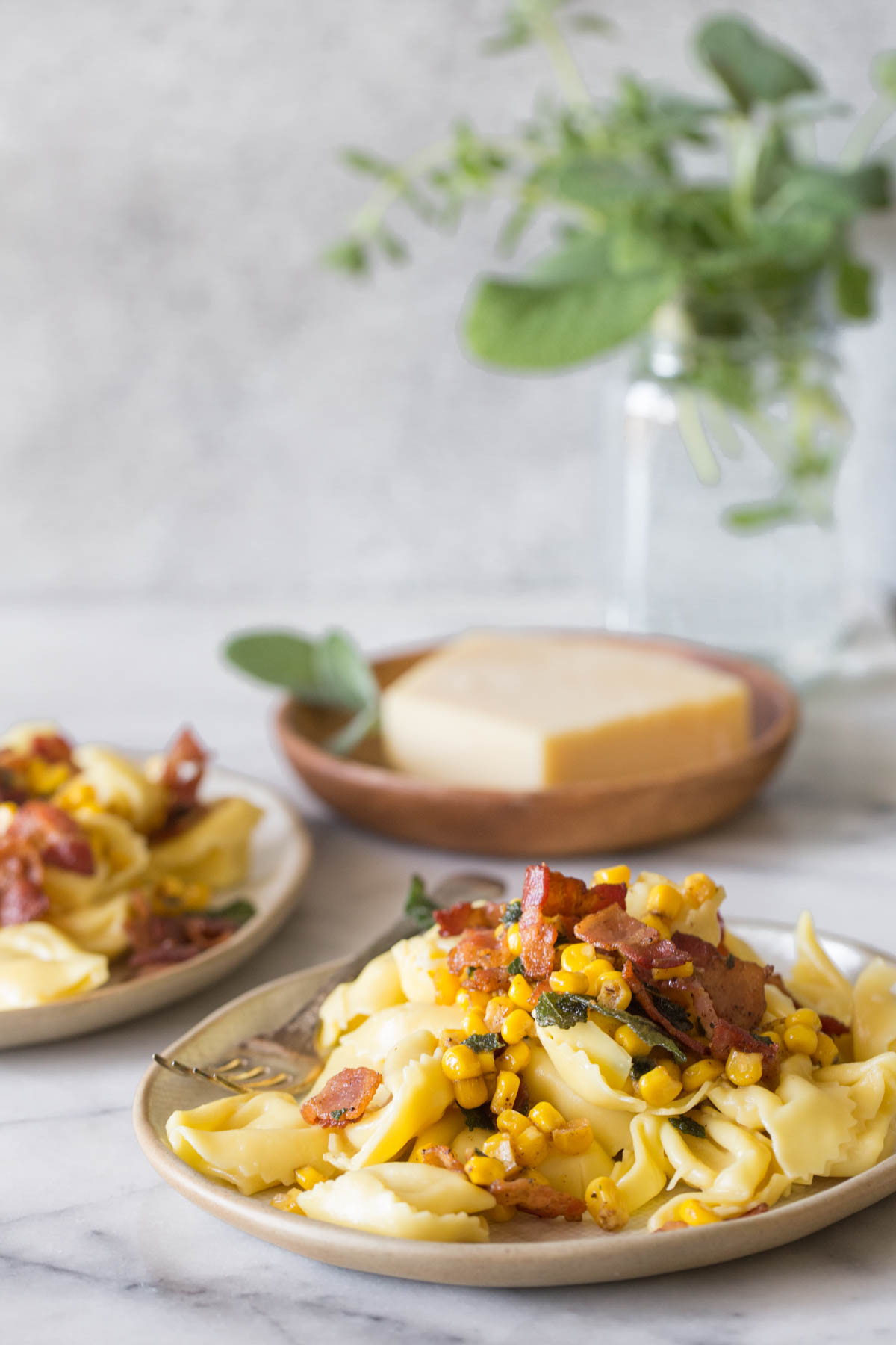 A plate of Creamy Tortellini With Bacon and Corn, and a wooden plate with a block of Parmesan cheese in the background.