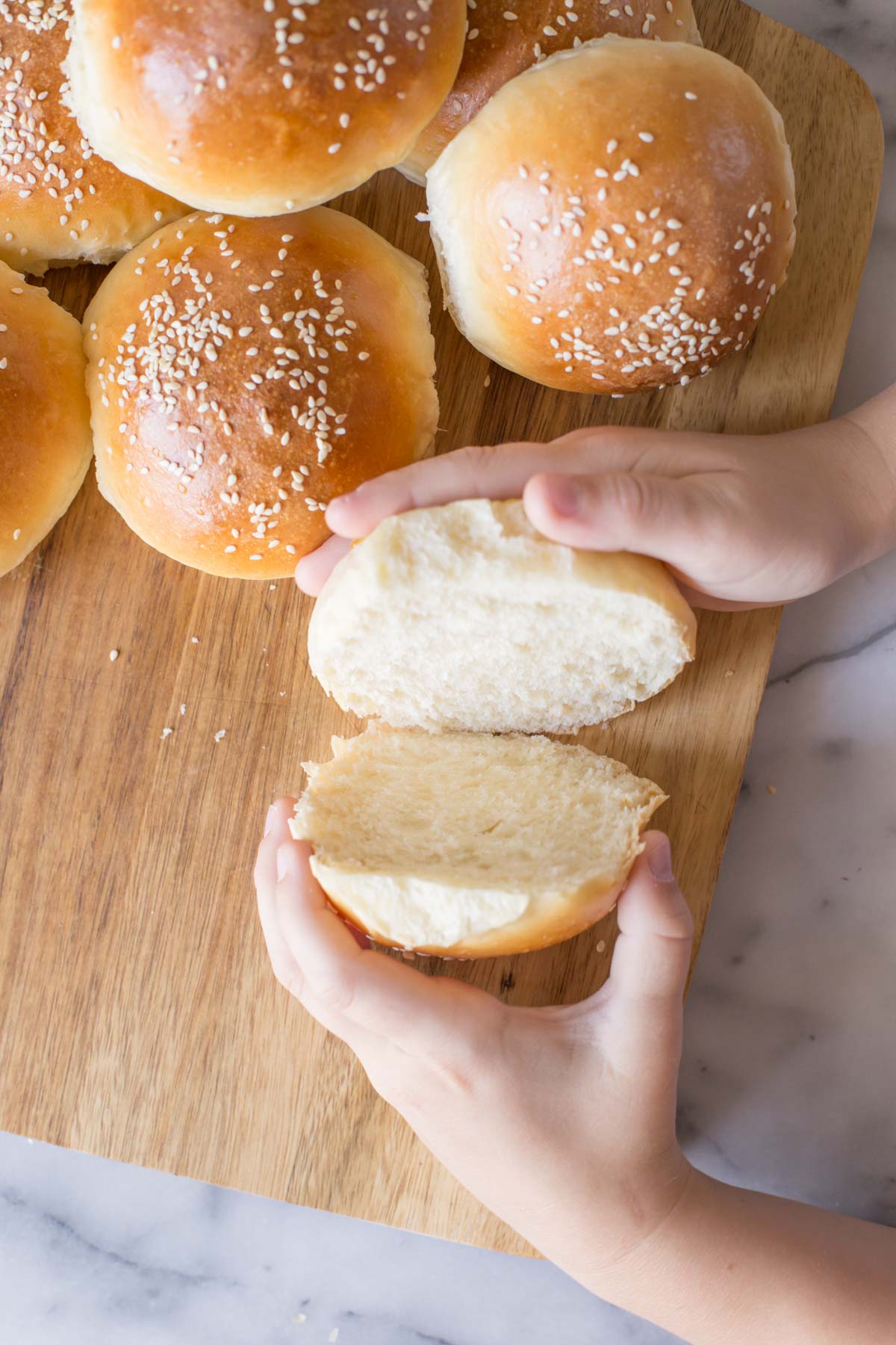 Two hands separating a cut Sourdough Hamburger Bun on a wooden cutting board with more buns next to it.  