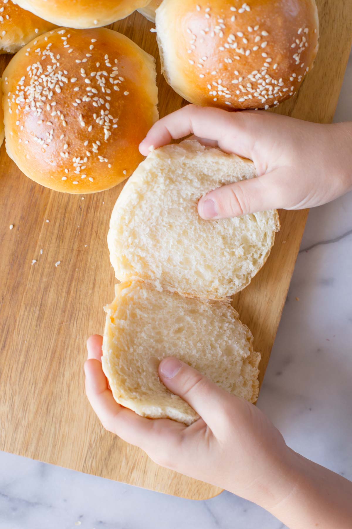 A close up shot of two hands separating a cut Sourdough Hamburger Bun on a wooden cutting board with more buns next to it.  