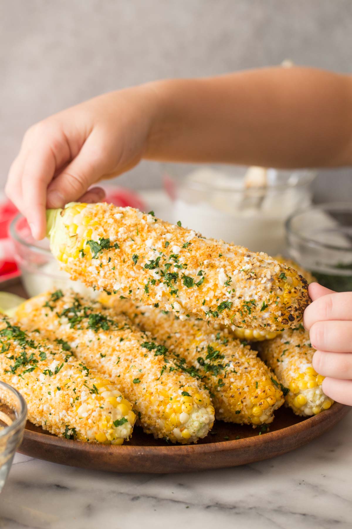Two hands holding an ear of Mexican Street Corn With Panko, with more ears of corn on a wood platter in the background.