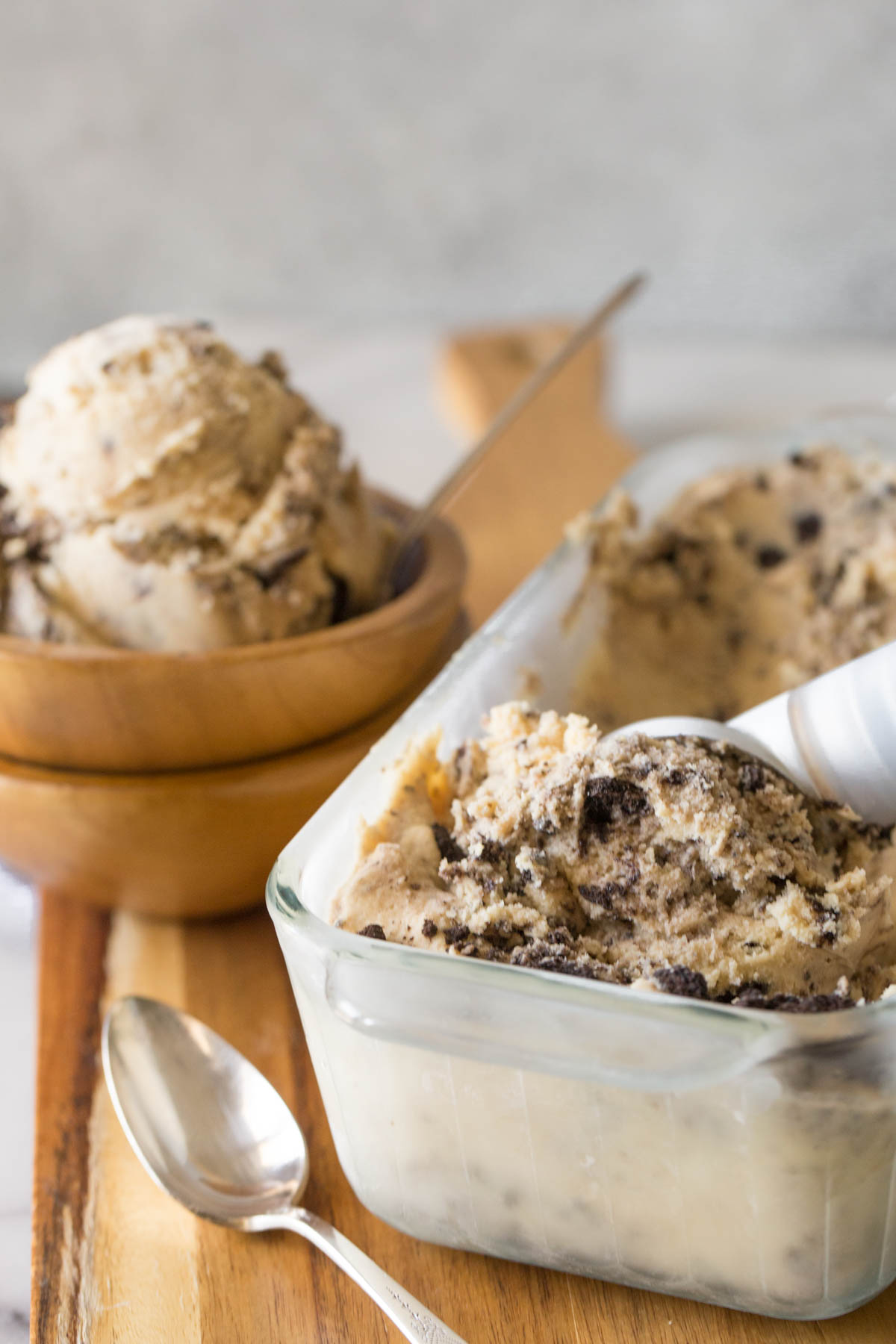 An ice cream scoop in a glass container of Peanut Butter Oreo Ice Cream, with a wood bowl of ice cream in the background.  