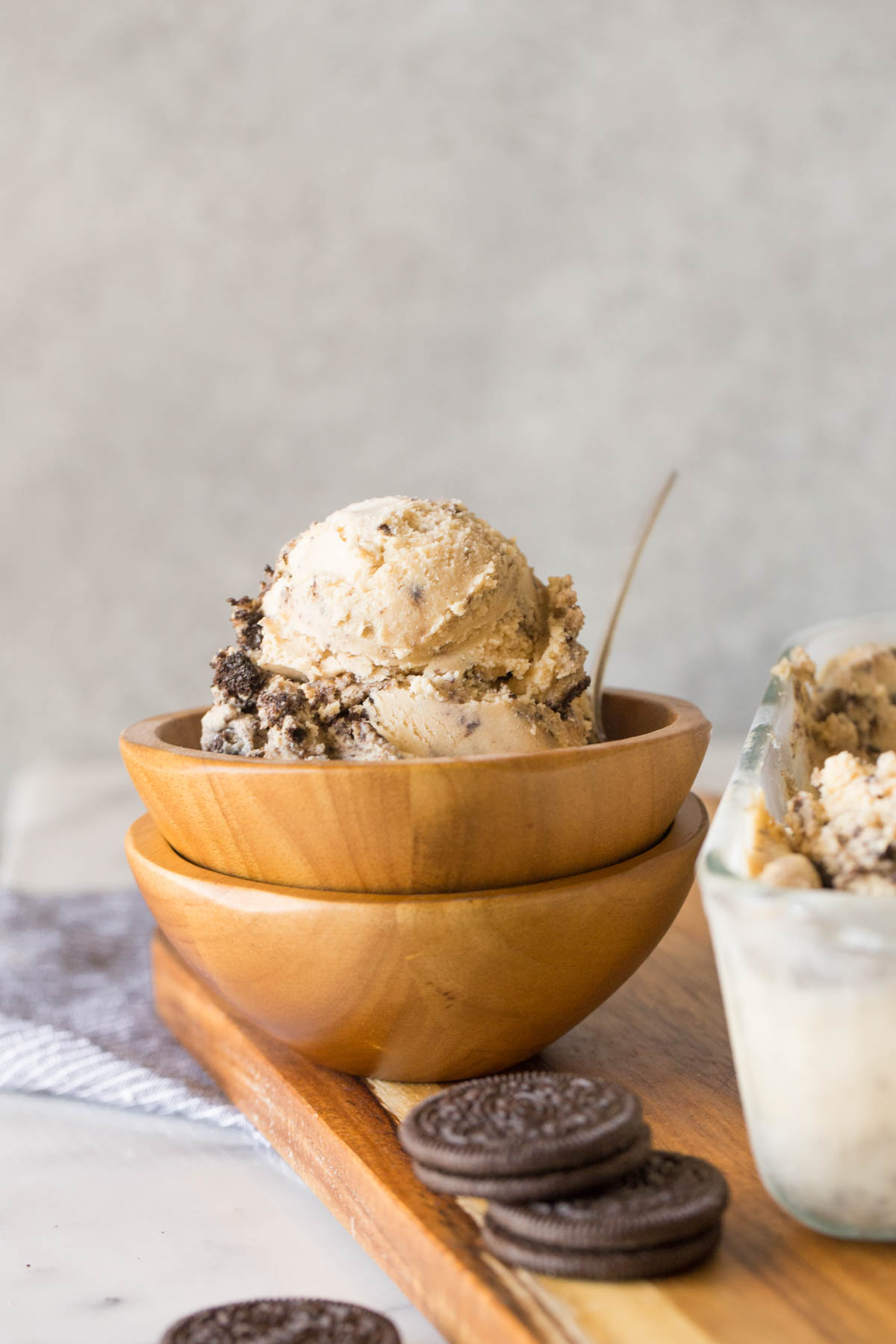 Peanut Butter Oreo Ice Cream in a wood bowl that is stacked on another wood bowl, with a few Oreo Cookies next to it along with a glass container of Peanut Butter Oreo Ice Cream.