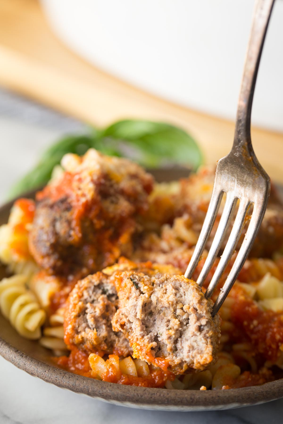 Close up shot of a fork in half of an Easy Broiled Italian Meatball, showing the inside texture, with a bowl of meatballs served over rotini pasta in the background.