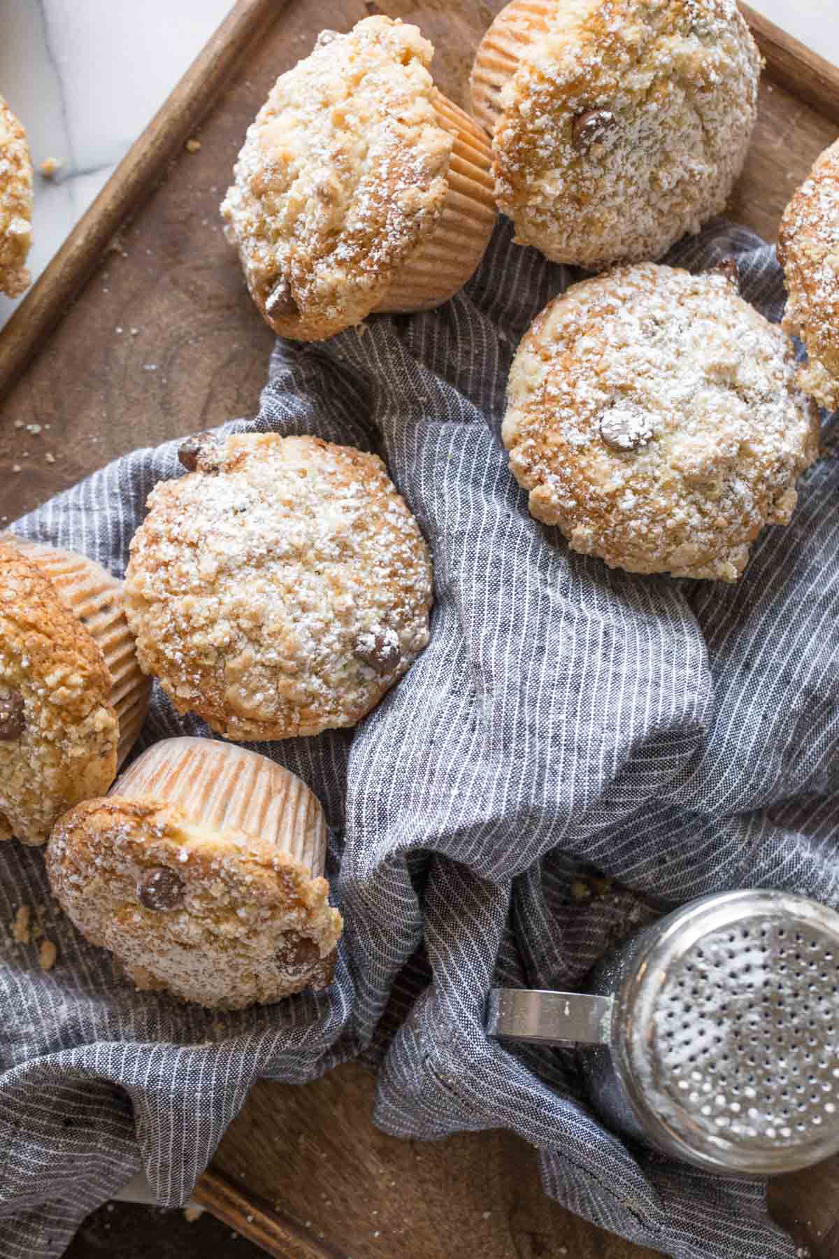 Overhead shot of Chocolate Chip Muffins and a powdered sugar shaker on a blue cloth on top of a wood cutting board. 