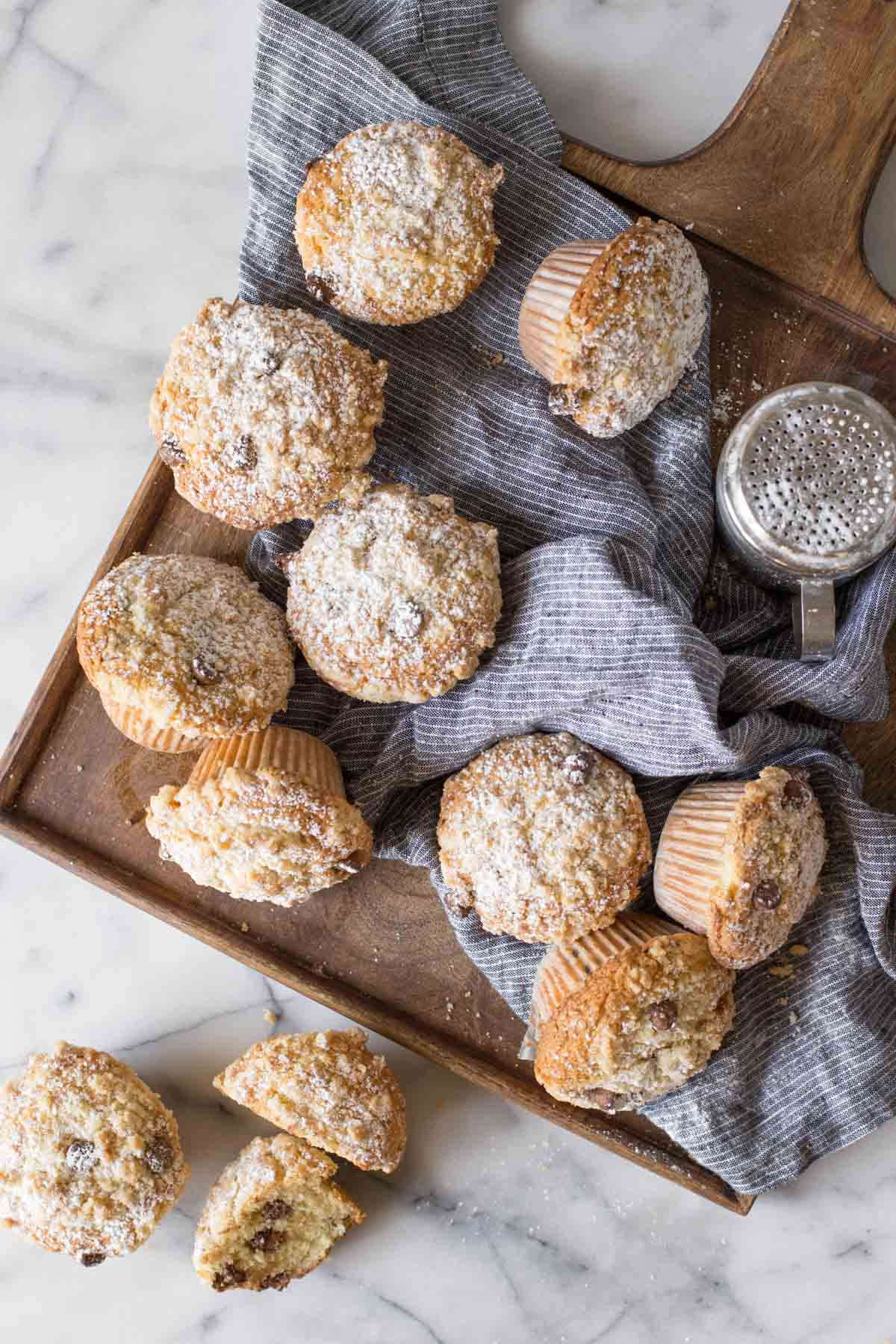 Overhead shot of Chocolate Chip Muffins and a powdered sugar shaker on a blue cloth on top of a wood cutting board with a marble background.  
