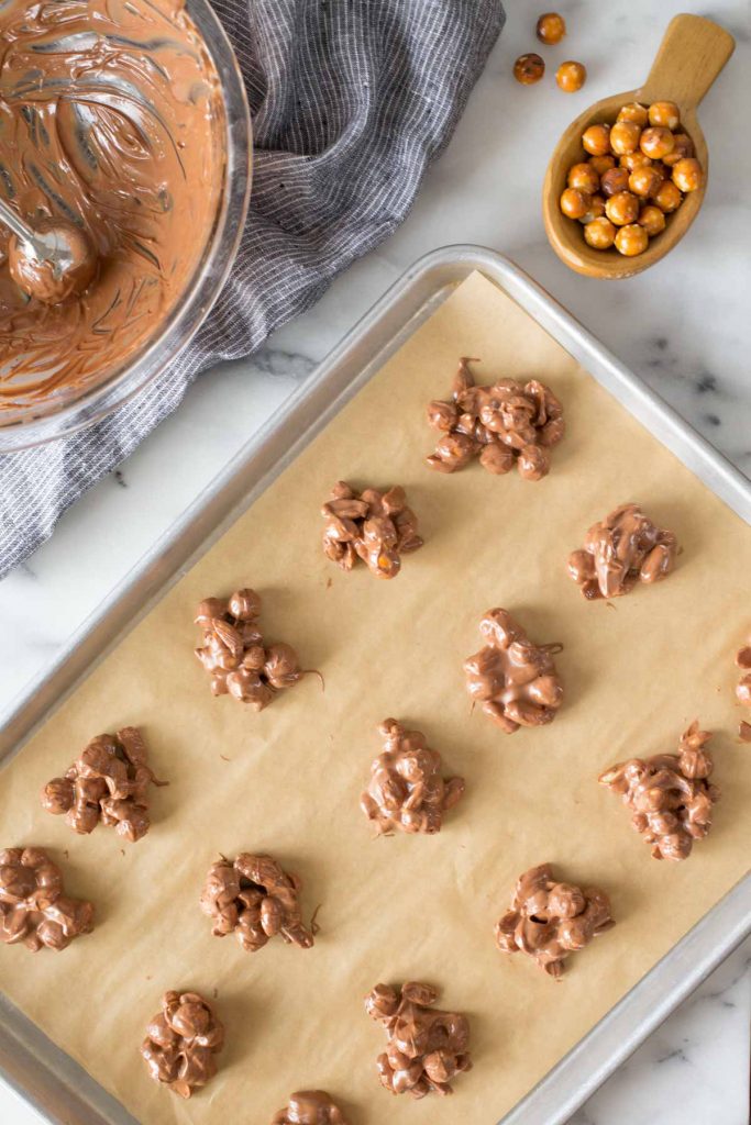Overhead shot of Chocolate Peanut Clusters on a parchment lined baking sheet, with the mixing bowl and a wooden spoon full of pretzel rounds next to the baking sheet.
