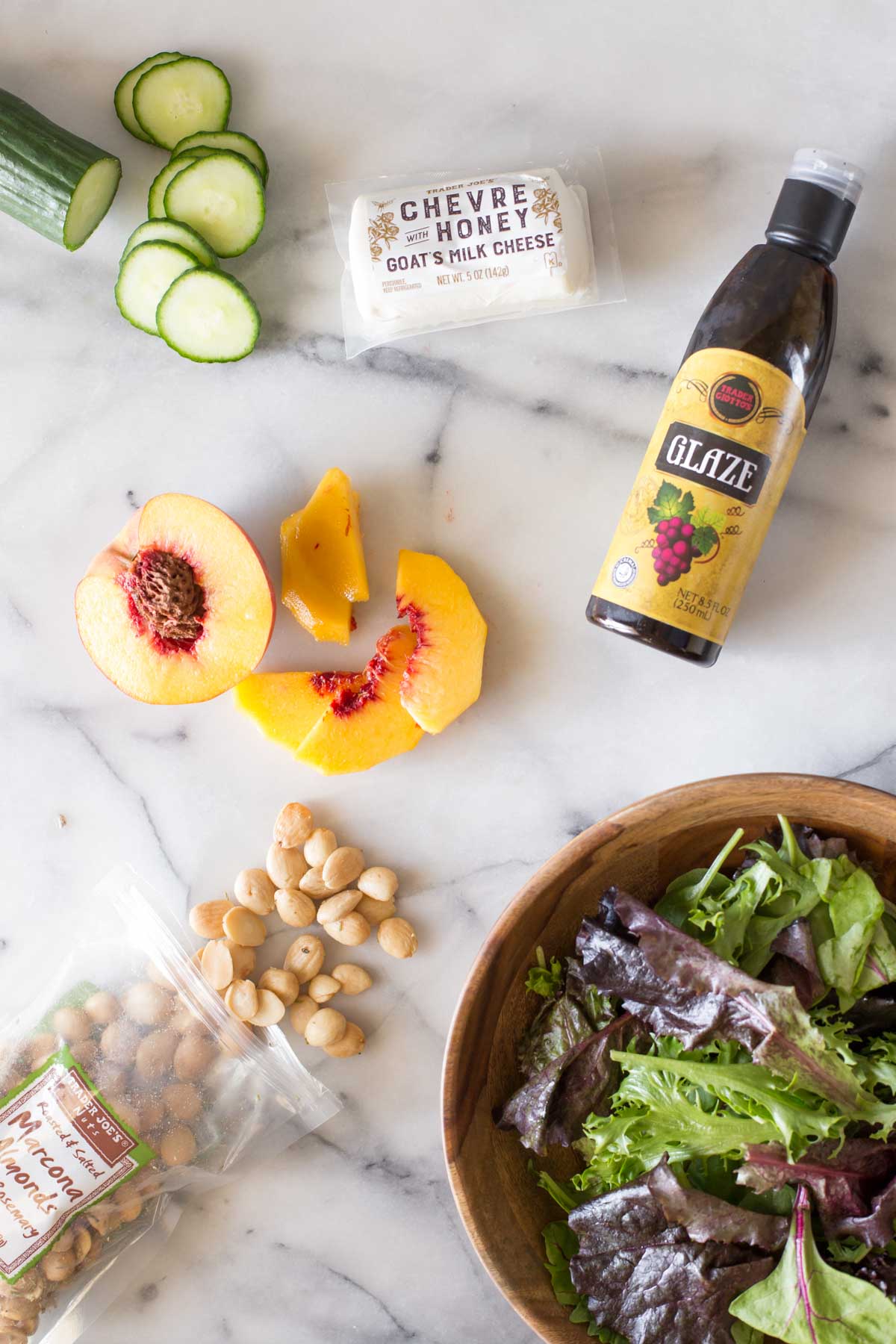 Overhead shot of a sliced cucumber, goat's milk cheese, a bottle of balsamic glaze, a sliced peach, roasted almonds, and a wood bowl of leafy greens on a marble background.