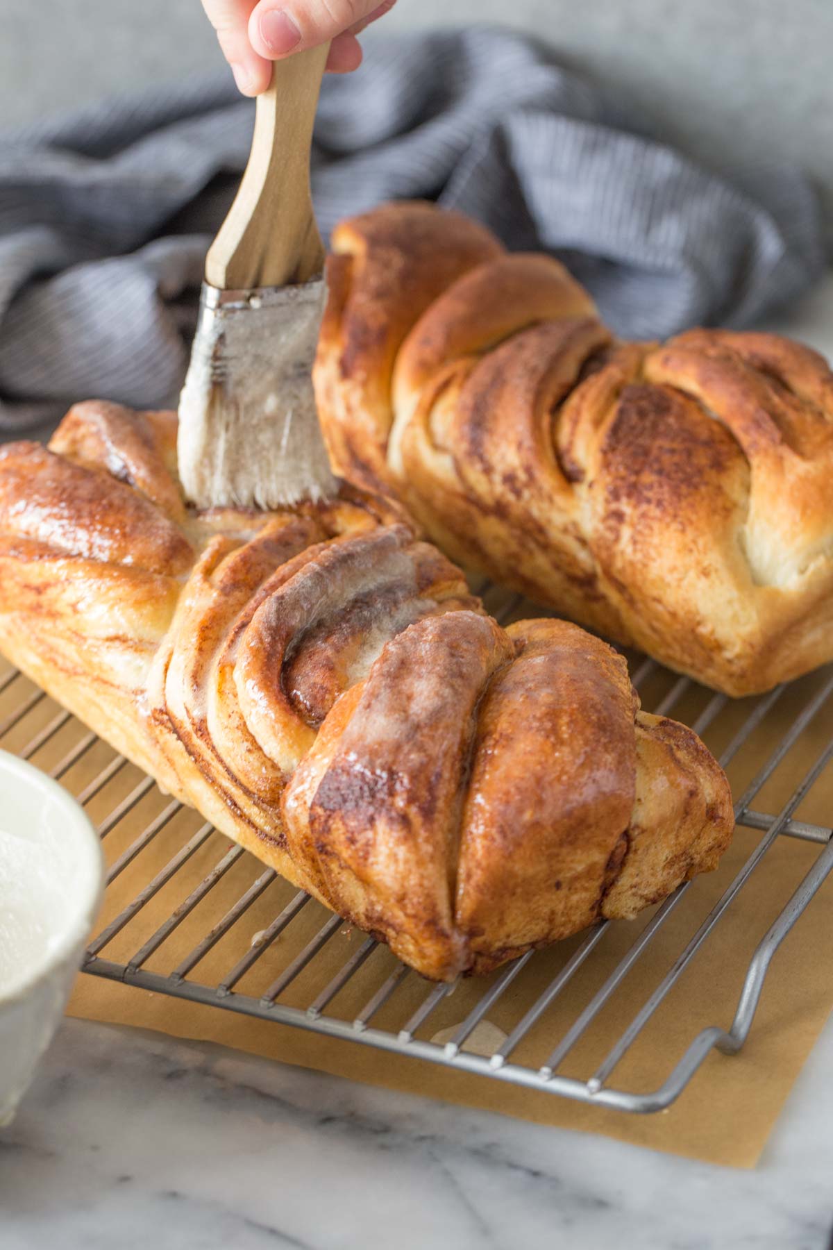 Icing being brushed onto a loaf of Cinnamon Twist Bread that is on a cooling rack with another loaf of Cinnamon Twist Bread, with parchment paper under the rack.