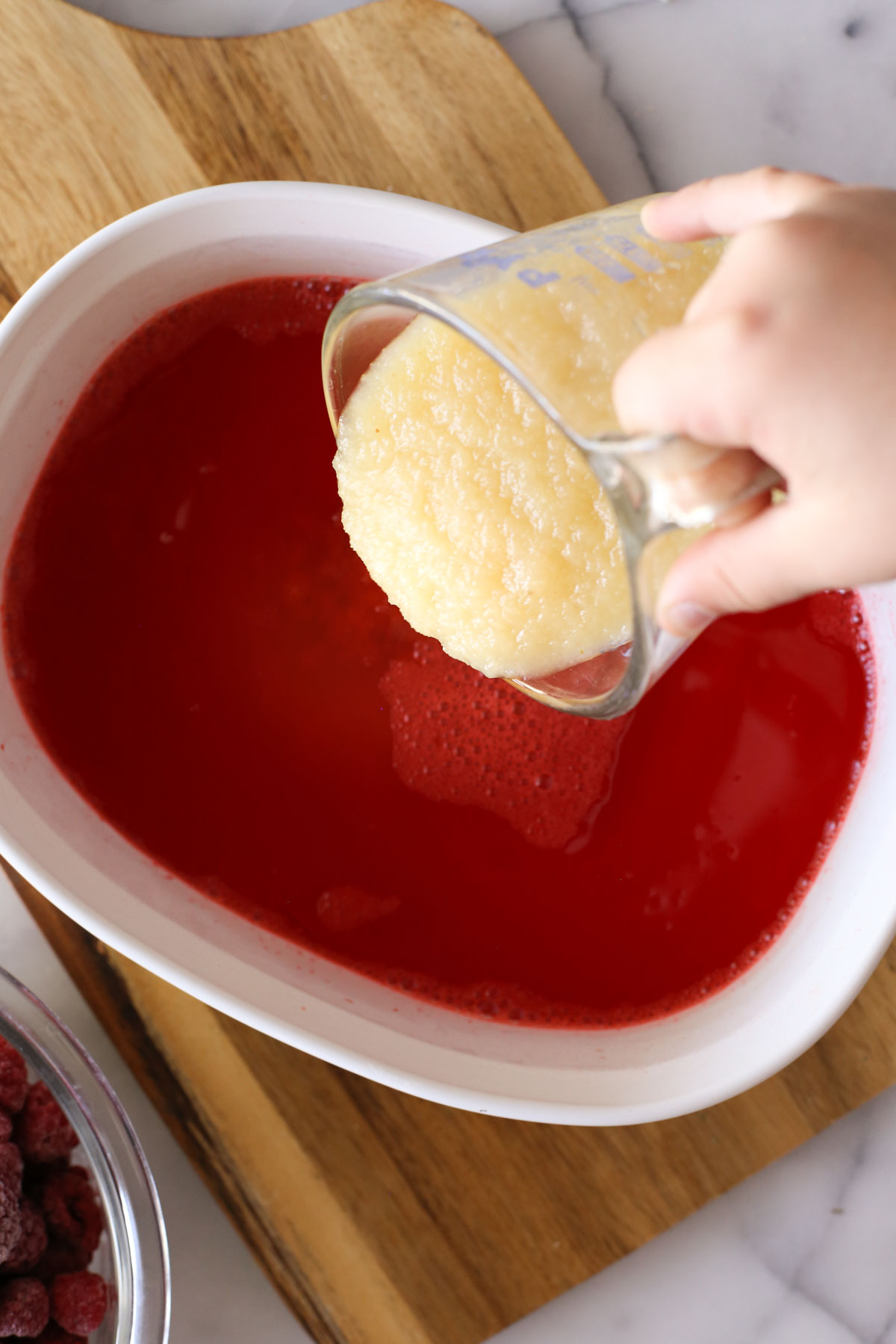 Overhead shot of a glass measuring cup of applesauce being poured into a baking dish of raspberry gelatin and boiling water.