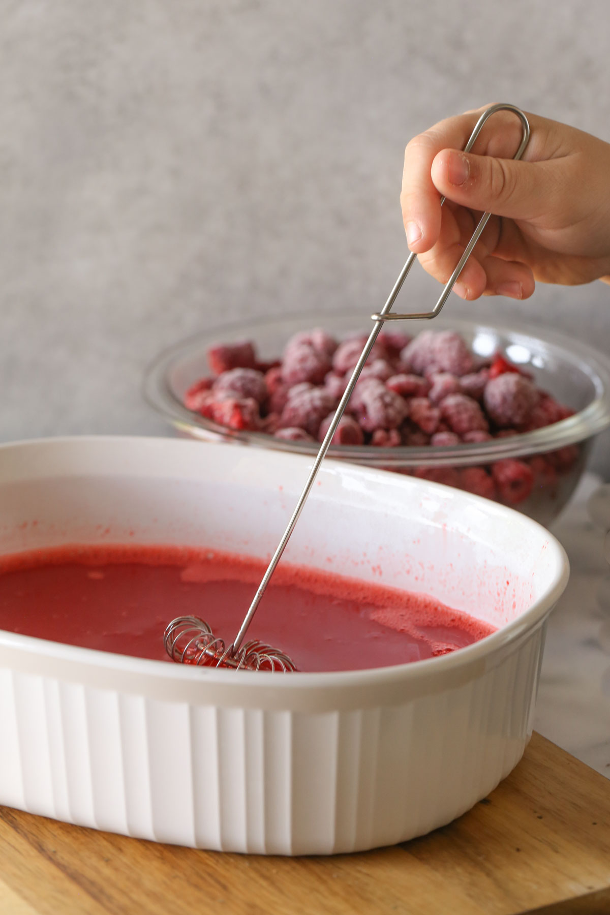 Ingredients for the Raspberry Jello Salad being whisked together in a baking dish, with a bowl of frozen raspberries in the background.