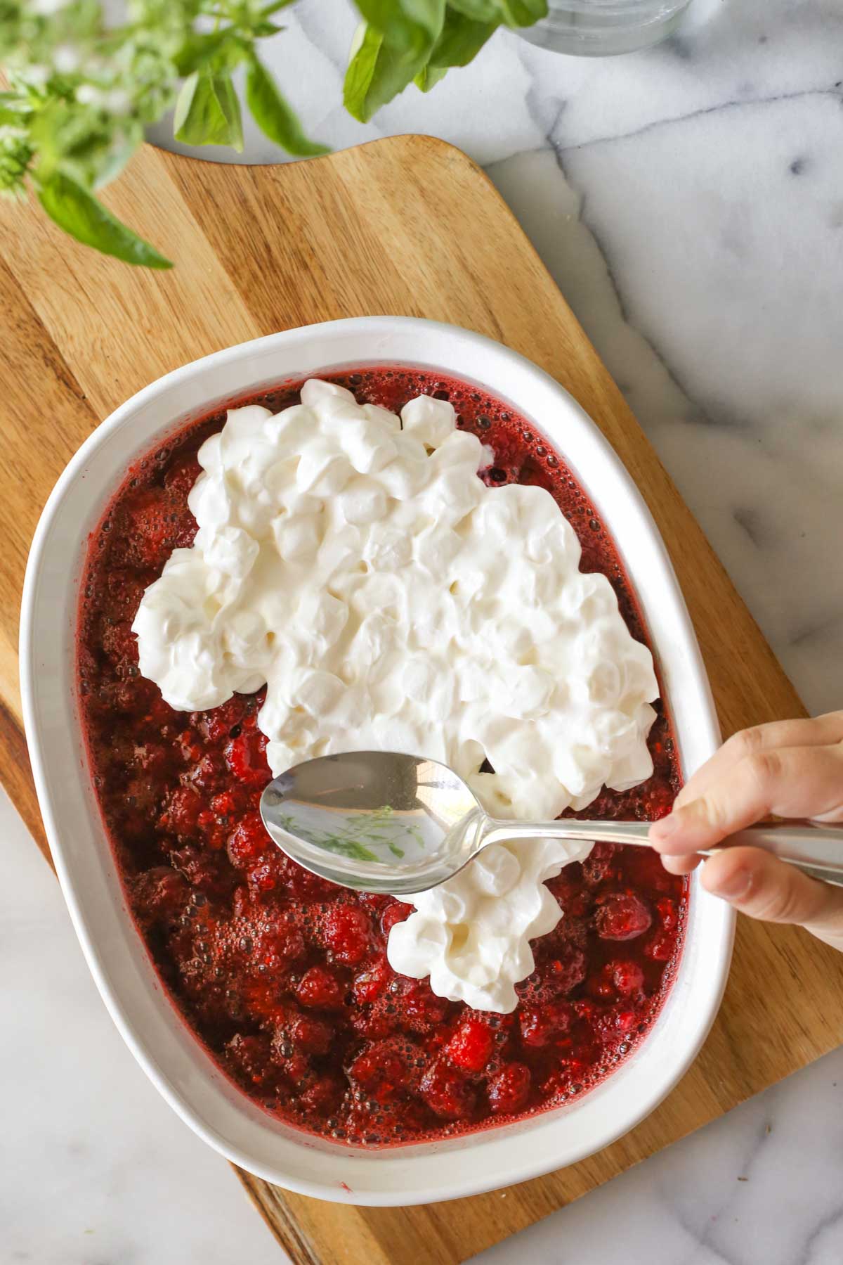 Overhead shot of Raspberry Jello Salad in a baking dish sitting on a wood cutting board, with the sour cream marshmallow mixture being spread on top.