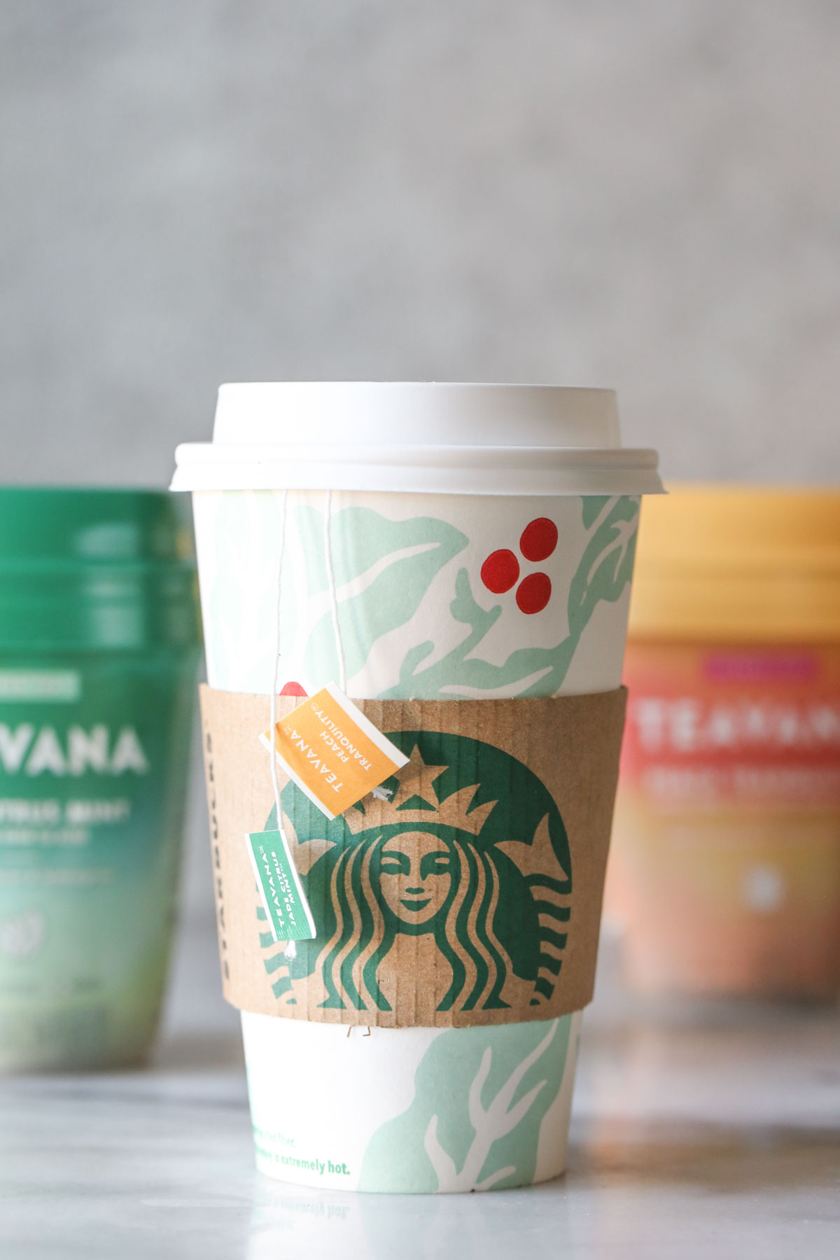 A Starbucks Medicine Ball Tea in a Starbucks hot cup, with the tea bag labels hanging over the side, and the Teavana tea packages in the background.