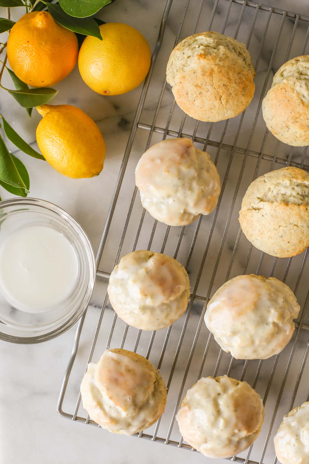 Overhead shot of Lemon Poppy Seed Muffins on a cooling rack, some glazed and some not yet glazed, with a glass bowl of glaze next to the cooling rack, along with a branch from a lemon tree and three whole lemons.