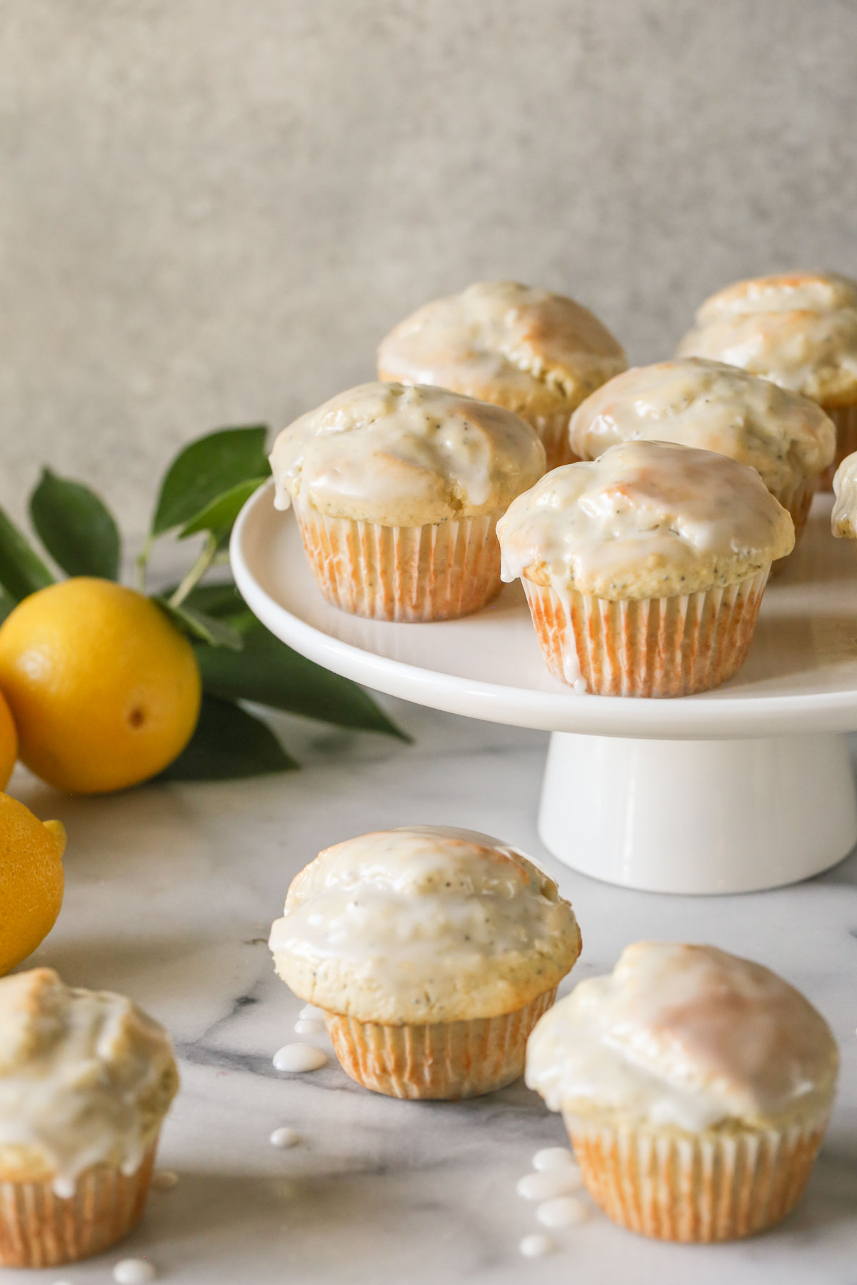 Glazed Lemon Poppy Seed Muffins on a cake stand with three muffins next to the stand, along with a branch from a lemon tree and whole lemons.