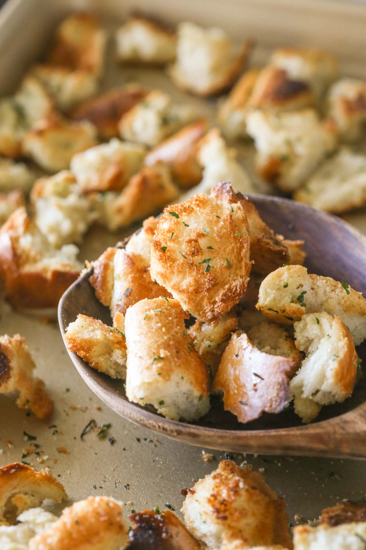 A wooden spoon full of Rustic Buttery Garlic Croutons being scooped off a baking sheet full of croutons.