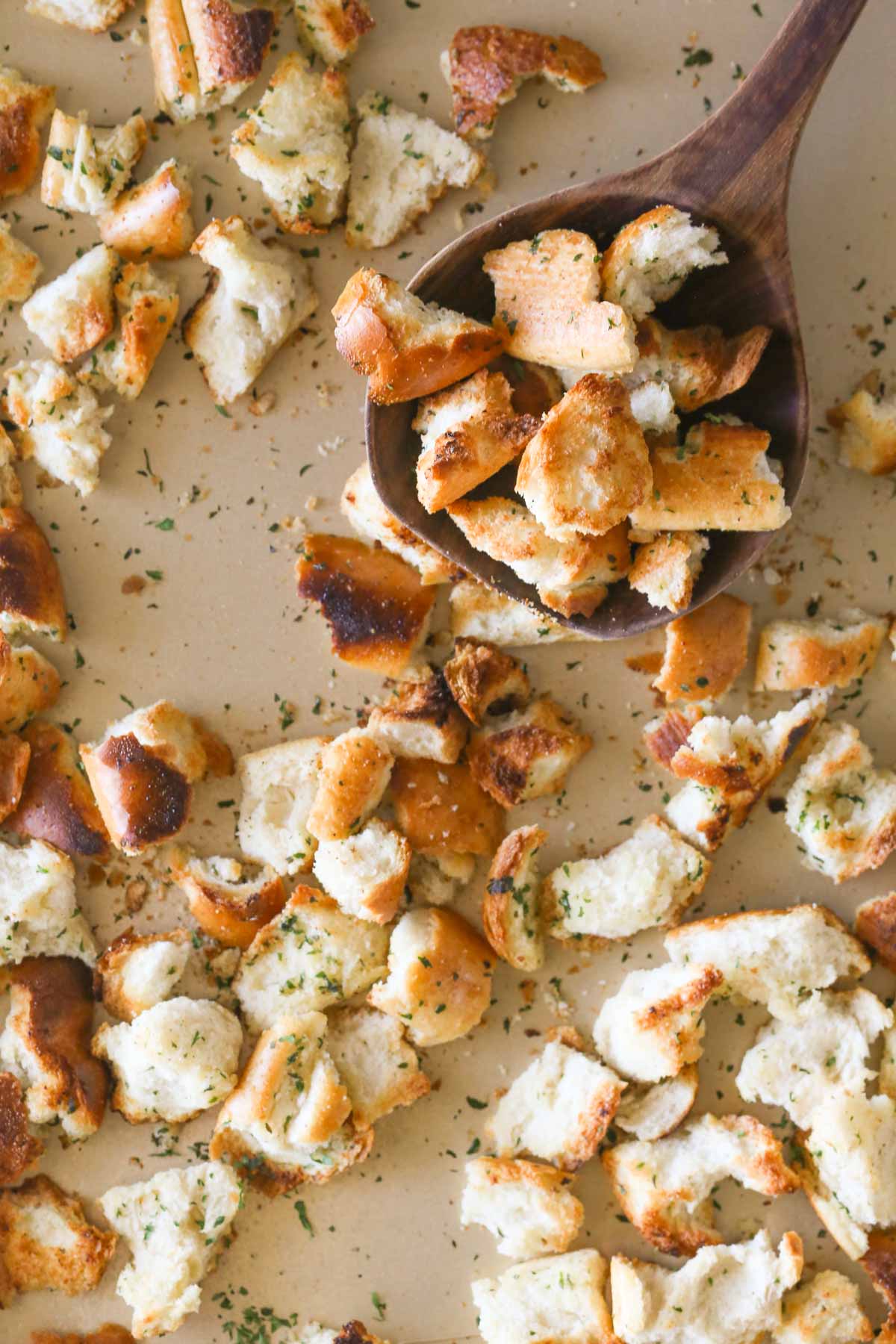 Overhead shot of a wooden spoon full of Rustic Buttery Garlic Croutons held over the baking sheet full of Rustic Buttery Garlic Croutons. 