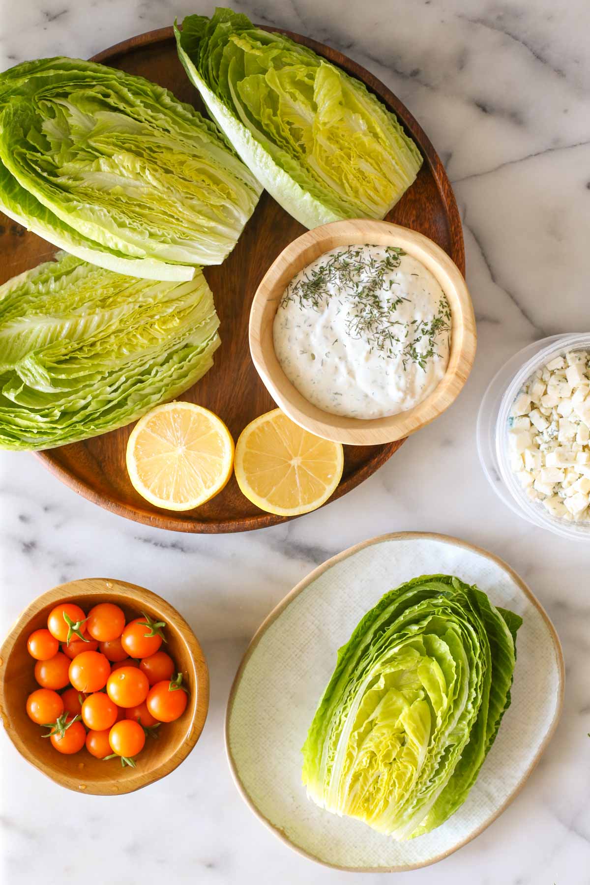 Overhead shot of a wood platter with three romaine lettuce halves, lemon wedges and a wood bowl of the blue cheese dressing on it, sitting next to a wood bowl of Sungold tomatoes, a container of blue cheese and a plate with a romaine lettuce half on it, all on a marble background.  