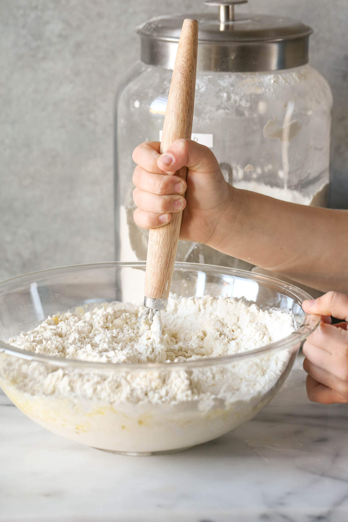 A mixing bowl with the ingredients for the Sourdough Sandwich Loaves in it, being mixed with a Danish Dough Whisk, with a large glass canister of flour in the background.