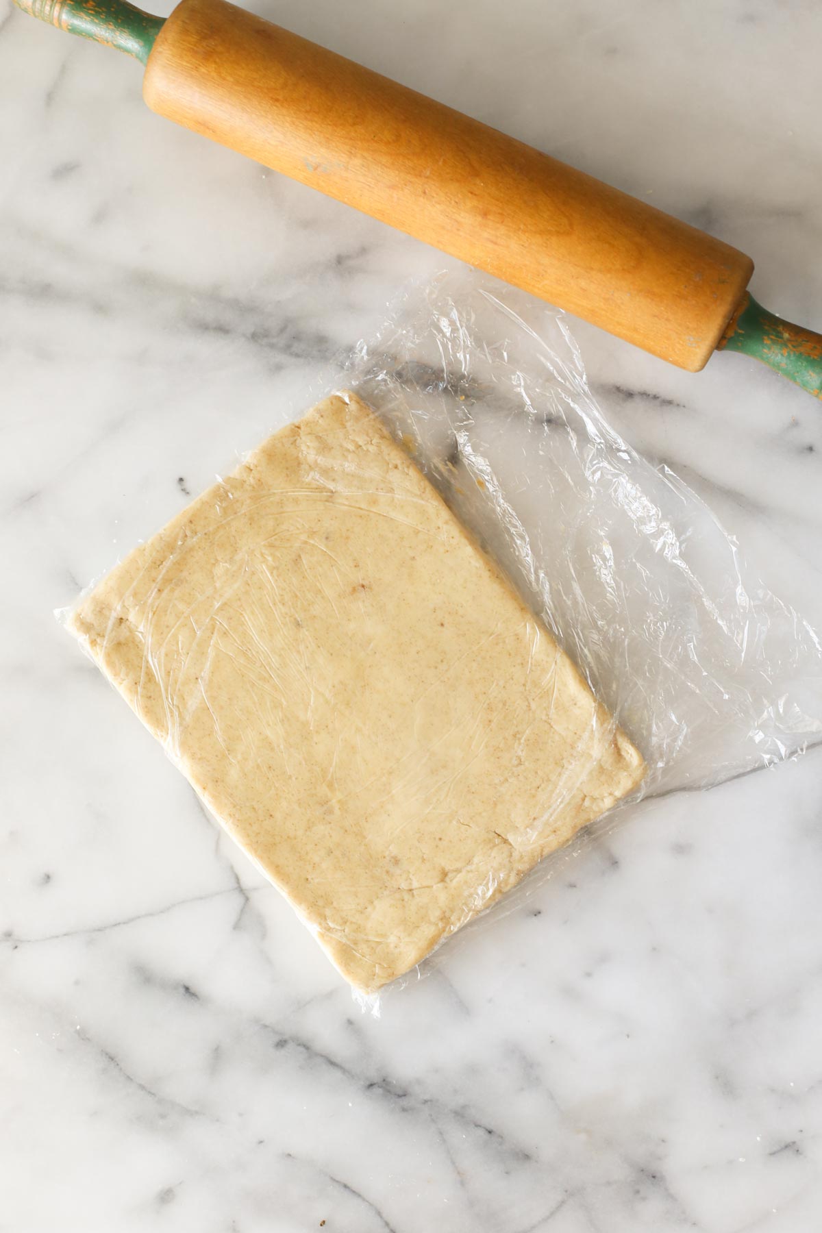 Overhead shot of the Buttery Shortbread Cookie dough in plastic wrap rolled out into a rectangle, with the rolling pin next to the dough, all on a marble background.