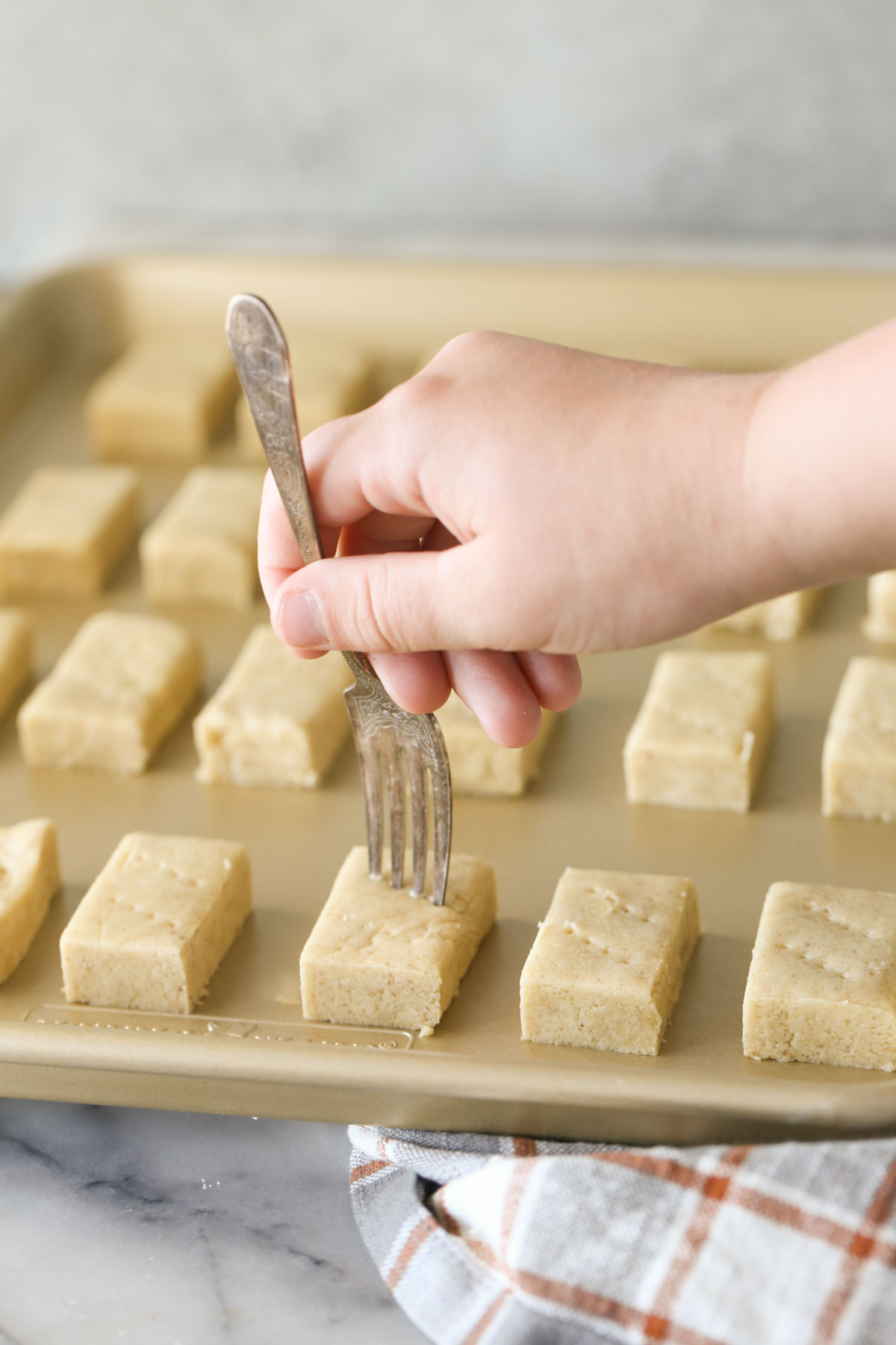 Buttery Shortbread Cookie dough rectangles arranged on a baking sheet, with a hand using a fork to poke the top of the dough.
