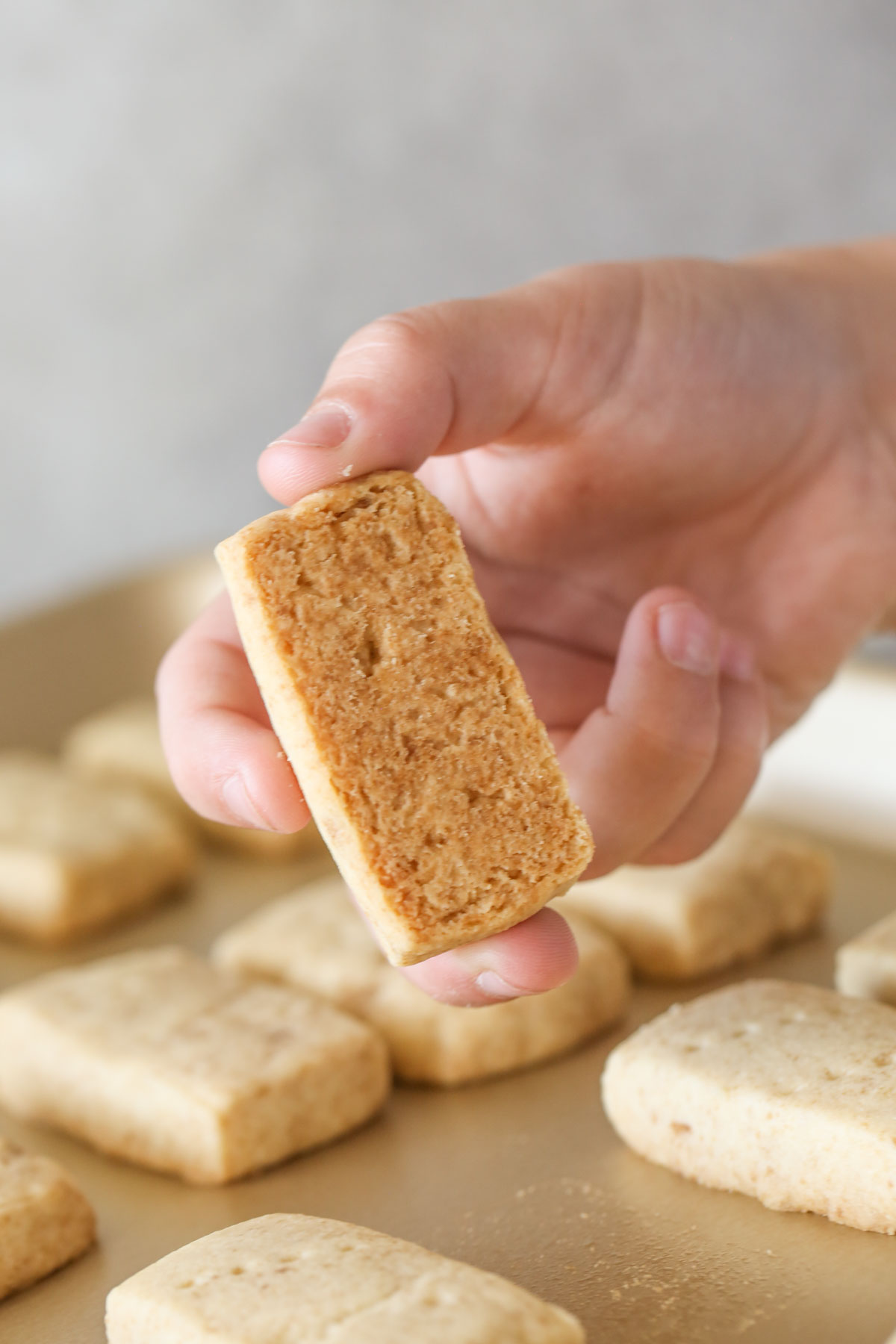 A hand holding a Buttery Shortbread Cookie showing the bottom of the baked cookie, held over the baking sheet with more Buttery Shortbread Cookies on it.