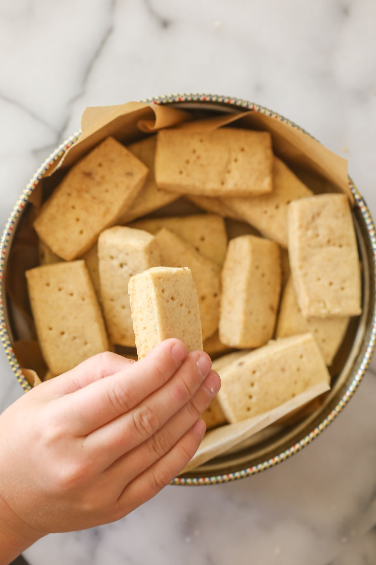 Overhead shot of a hand holding a Buttery Shortbread Cookie over a round cookie tin full of Buttery Shortbread Cookies.