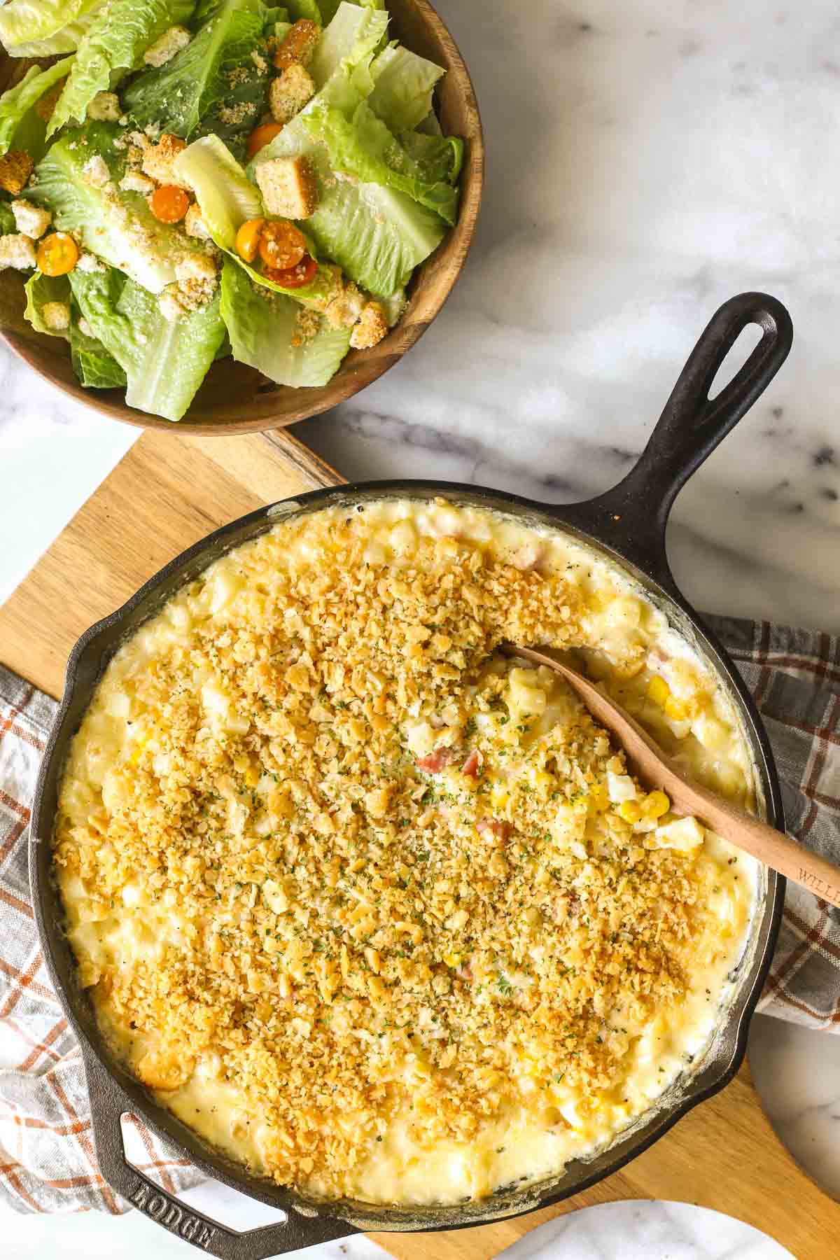 Overhead shot of Cheesy Ham and Potato Skillet in a cast iron skillet sitting on a wood cutting board, with a bowl of salad next to it, all on a marble background. 
