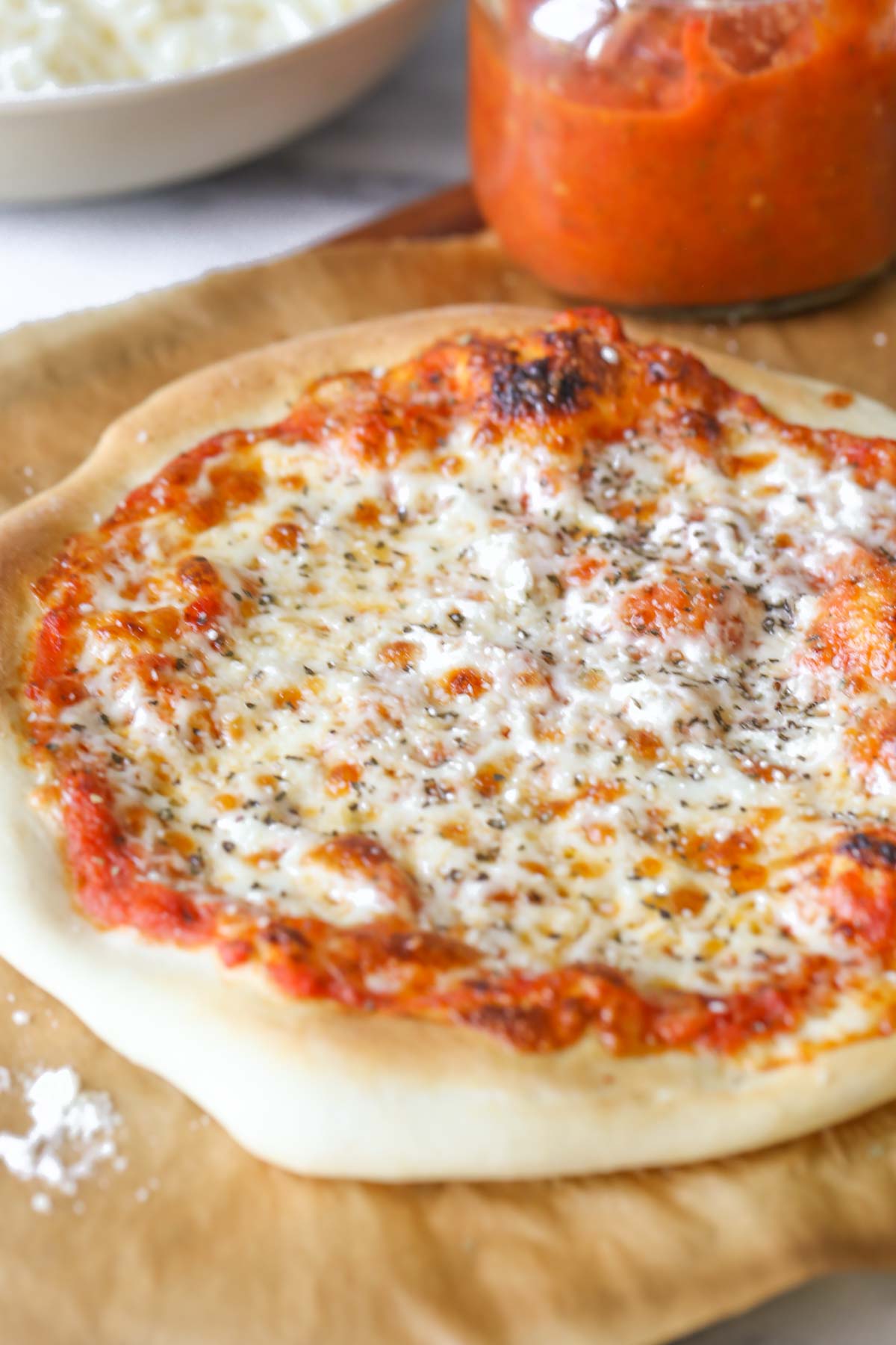 Close up shot of a homemade cheese pizza on parchment paper, sitting on a wood cutting board with a jar of Roasted Tomato Pizza Sauce.