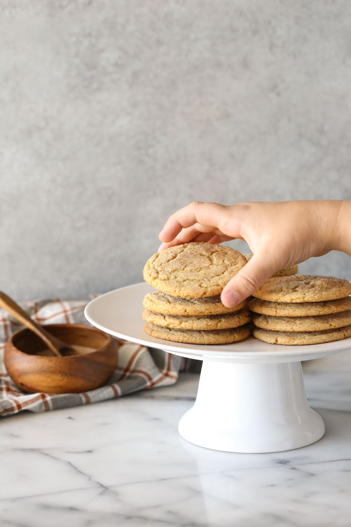 Brown Butter Snickerdoodles stacked on a white cake stand, with a hand picking up the cookie on top of one of the stacks, and a small wood bowl of cinnamon sugar in the background.