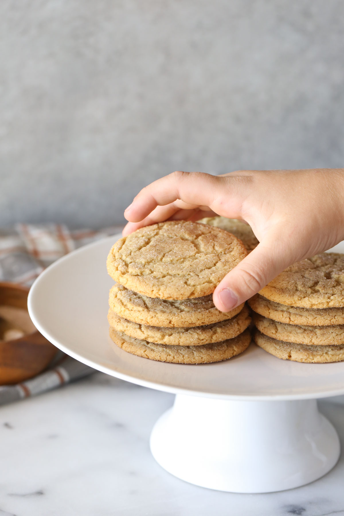 Close up shot of Brown Butter Snickerdoodles stacked on a white cake stand, with a hand picking up the cookie on top of one of the stacks.