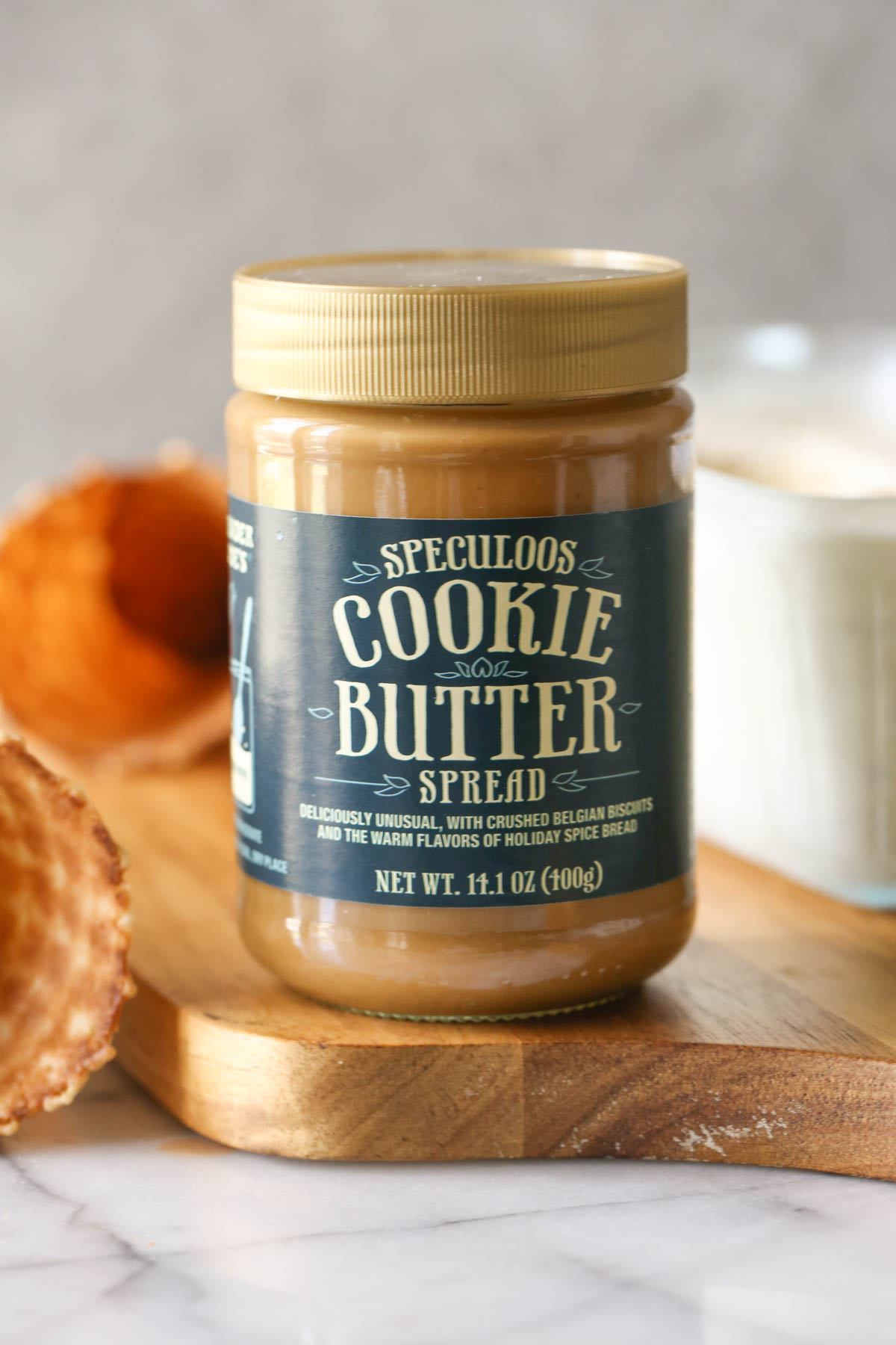 A jar of Speculoos Cooke Butter Spread on a wood cutting board, with waffle cones and a container of Cookie Butter Crisp Ice Cream in the background.
