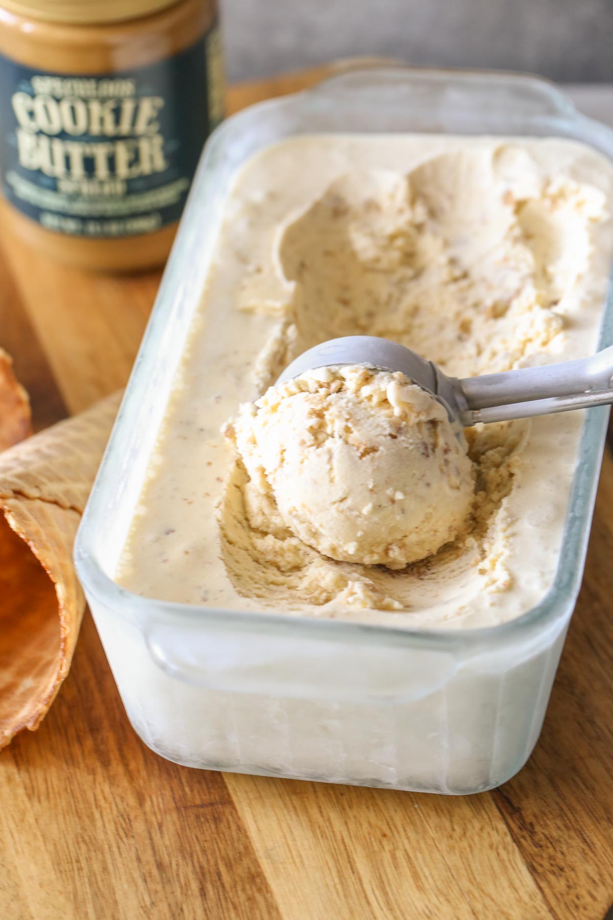 Cookie Butter Crisp Ice Cream in a glass container, with an ice cream scooper full of ice cream, and a waffle cone next to the container, along with a jar of cookie butter in the background.