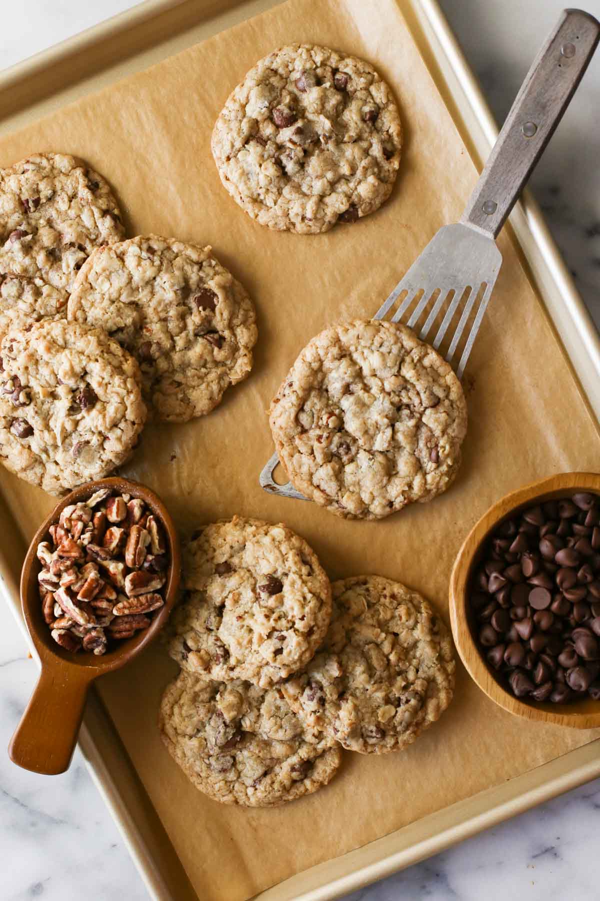 Overhead shot of Cowboy Cookies on a parchment paper lined baking sheet, along with a wood scoop full of pecans and a wood bowl of chocolate chips, and a spatula under one of the Cowboy Cookies.