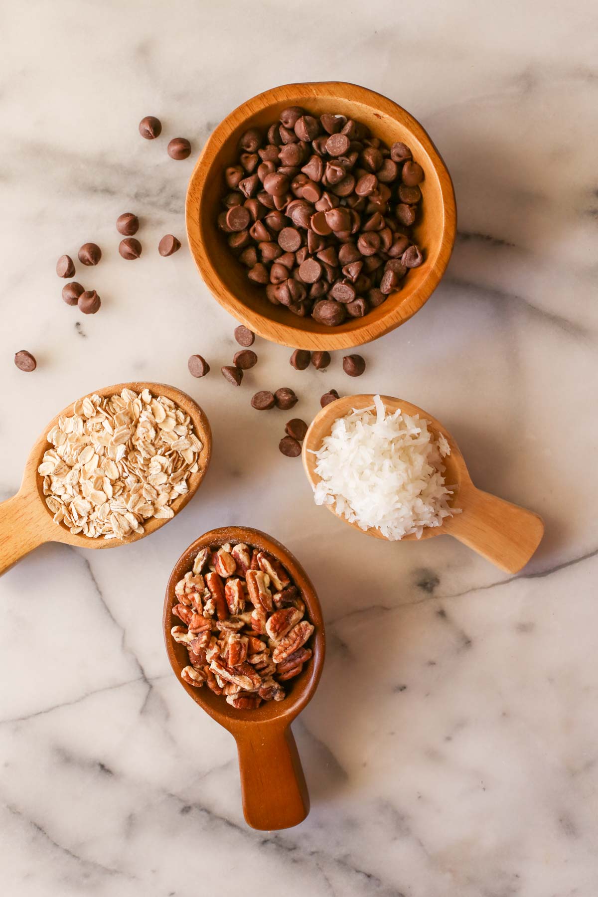 Overhead shot of a wood bowl of chocolate chips, with some of the chocolate chips next to the bowl, a small wood scoop of rolled oats, a small wood scoop of pecans, and a small wood scoop of shredded coconut, all on a marble background.