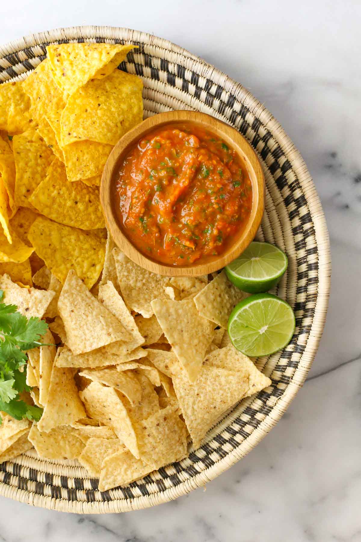 Overhead shot of a basket with tortilla chips, fresh cilantro, a lime cut in half, and a small wood bowl of Roasted Tomato Salsa.