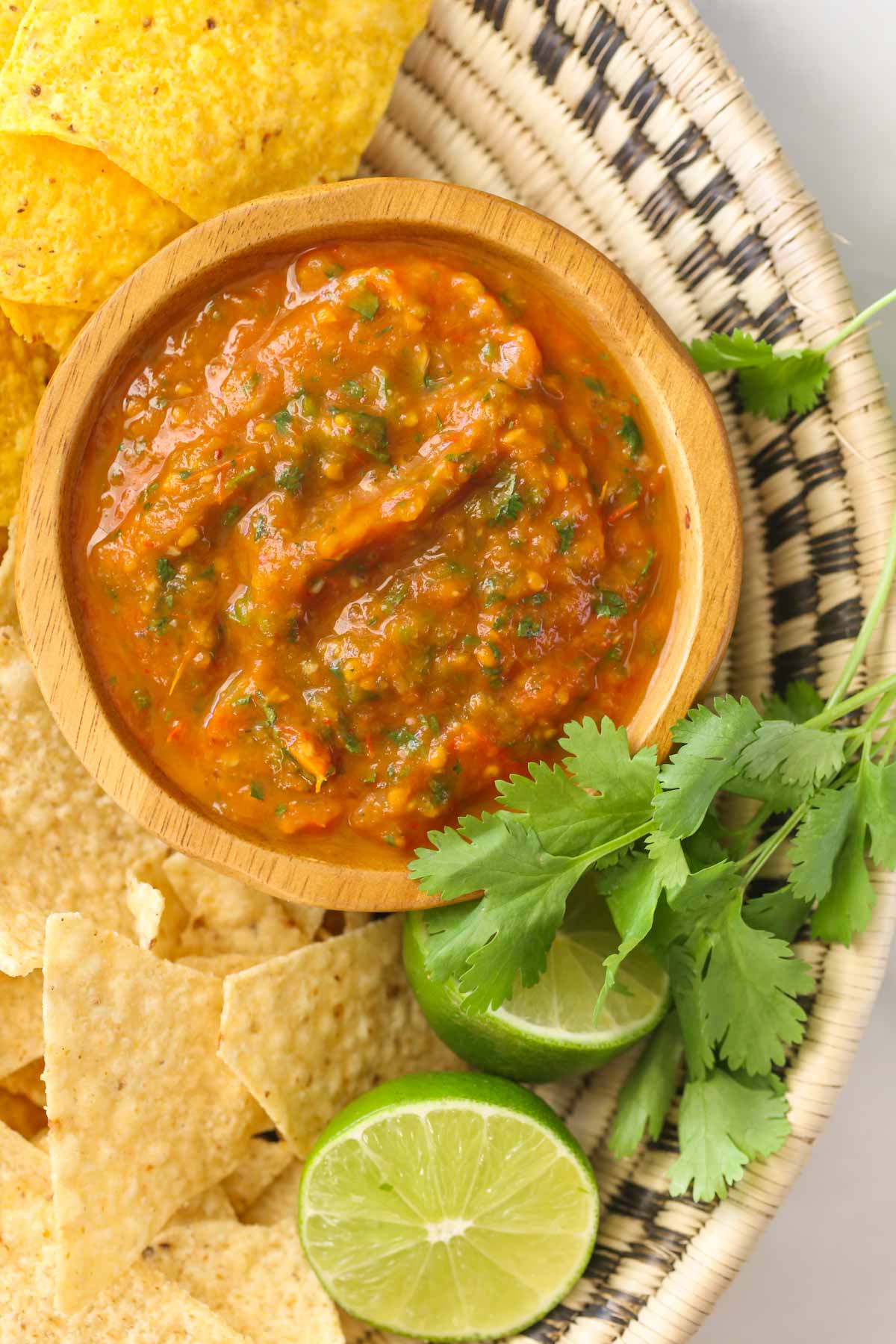 Close up shot of a small wood bowl of Roasted Tomato Salsa in a basket along with tortilla chips, fresh cilantro, and two lime halves.