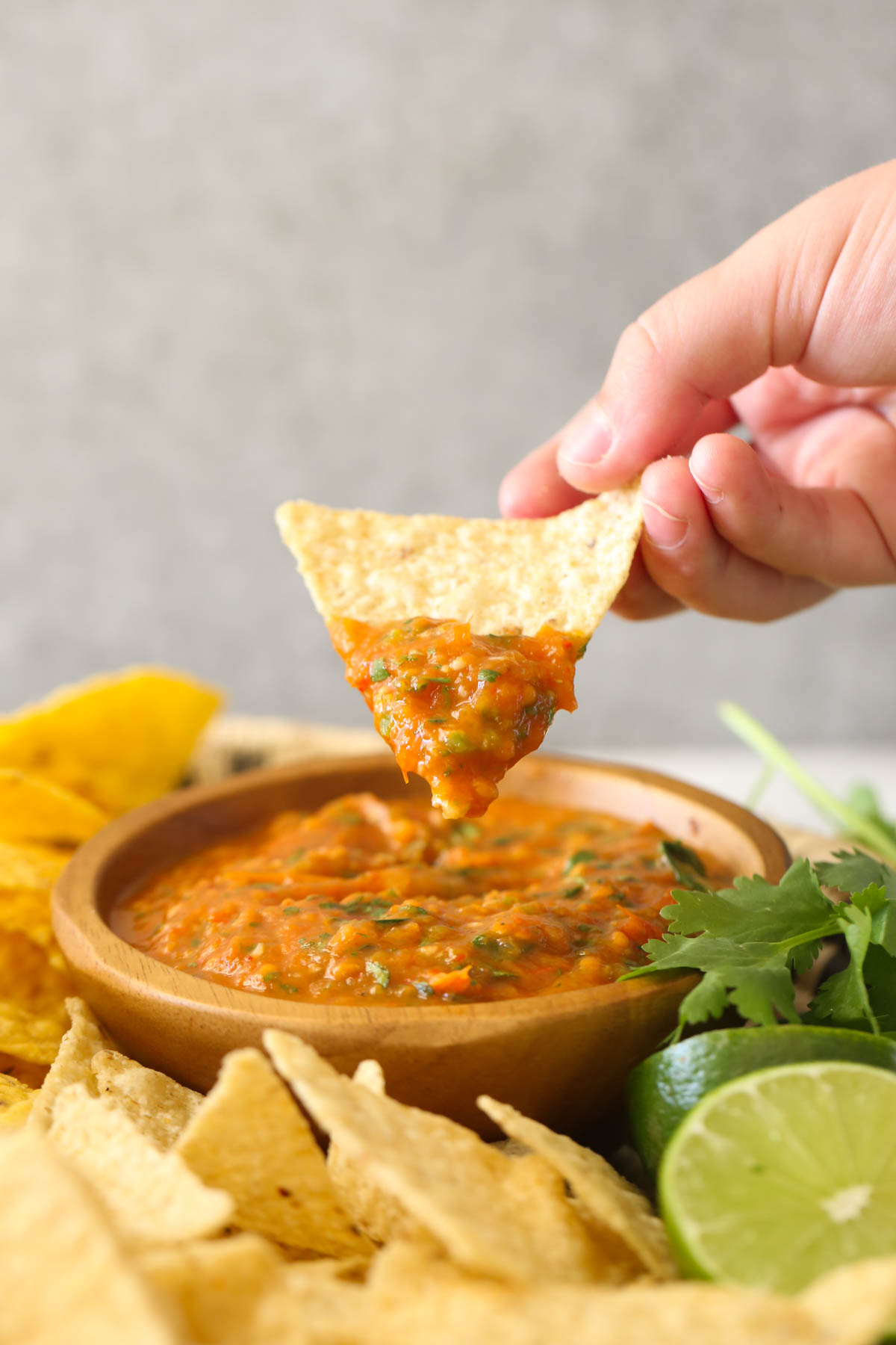 A hand holding a tortilla chip with Roasted Tomato Salsa on it above a small wood bowl of Roasted Tomato Salsa with tortilla chips, two lime halves and fresh cilantro next to the bowl.