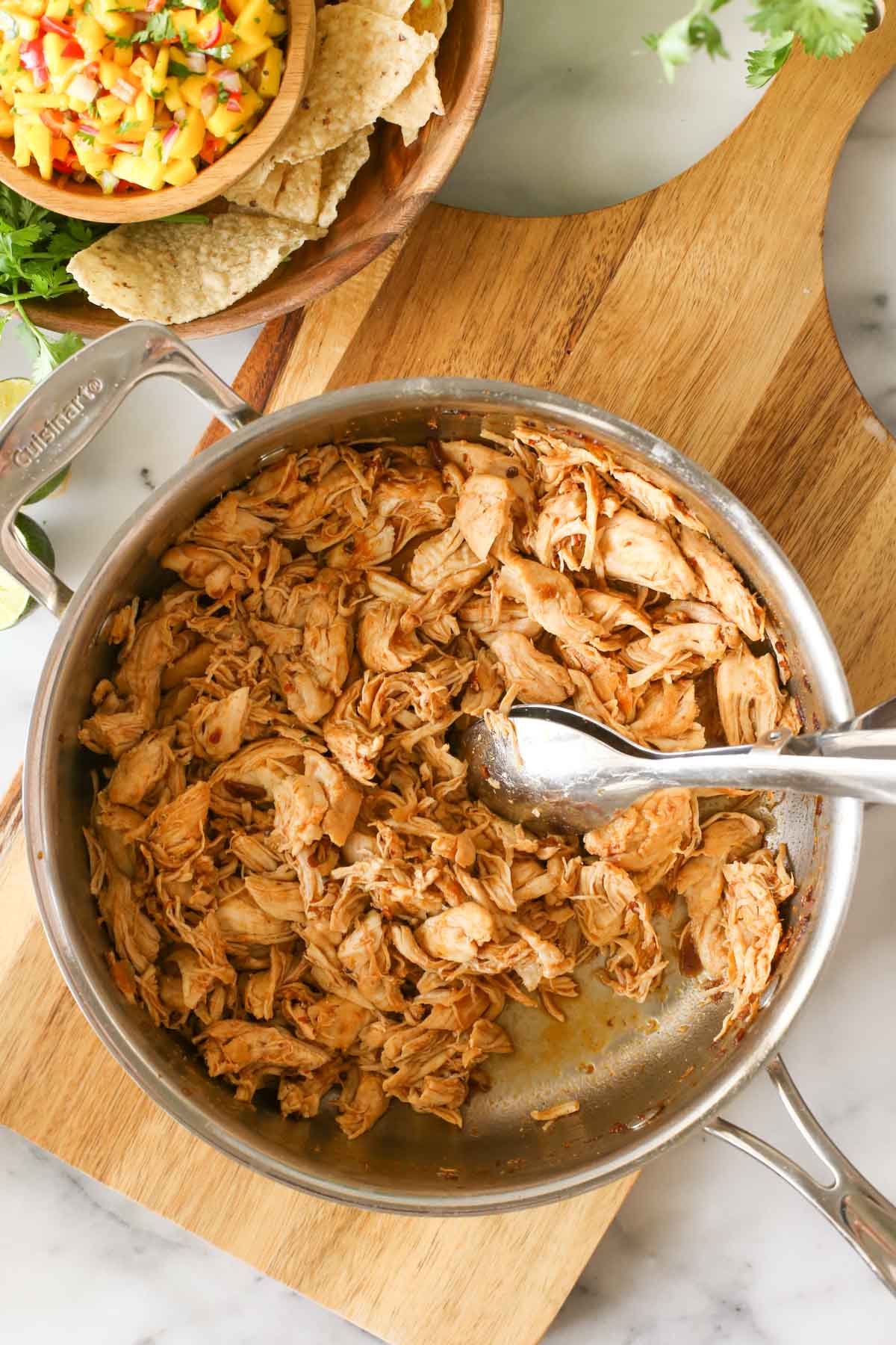 Overhead shot of a skillet of Buttery Honey Chipotle Chicken with a serving utensil in it, sitting on a wood cutting board next to a wood bowl of tortilla chips, fresh cilantro and a bowl of Mango Lime Salsa.
