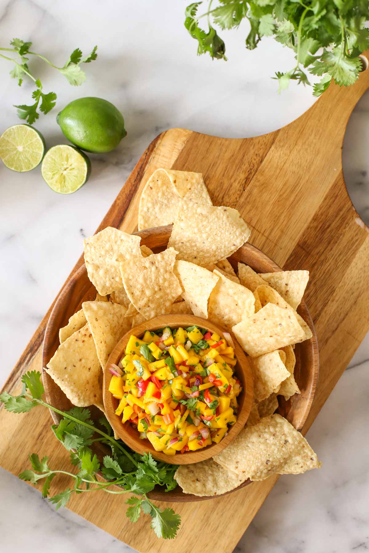 Overhead shot of a wood bowl of Mango Lime Salsa sitting inside a larger wood bowl with tortilla chips and fresh cilantro, all sitting on a wood cutting board with limes and cilantro next to it, all on a marble background. 