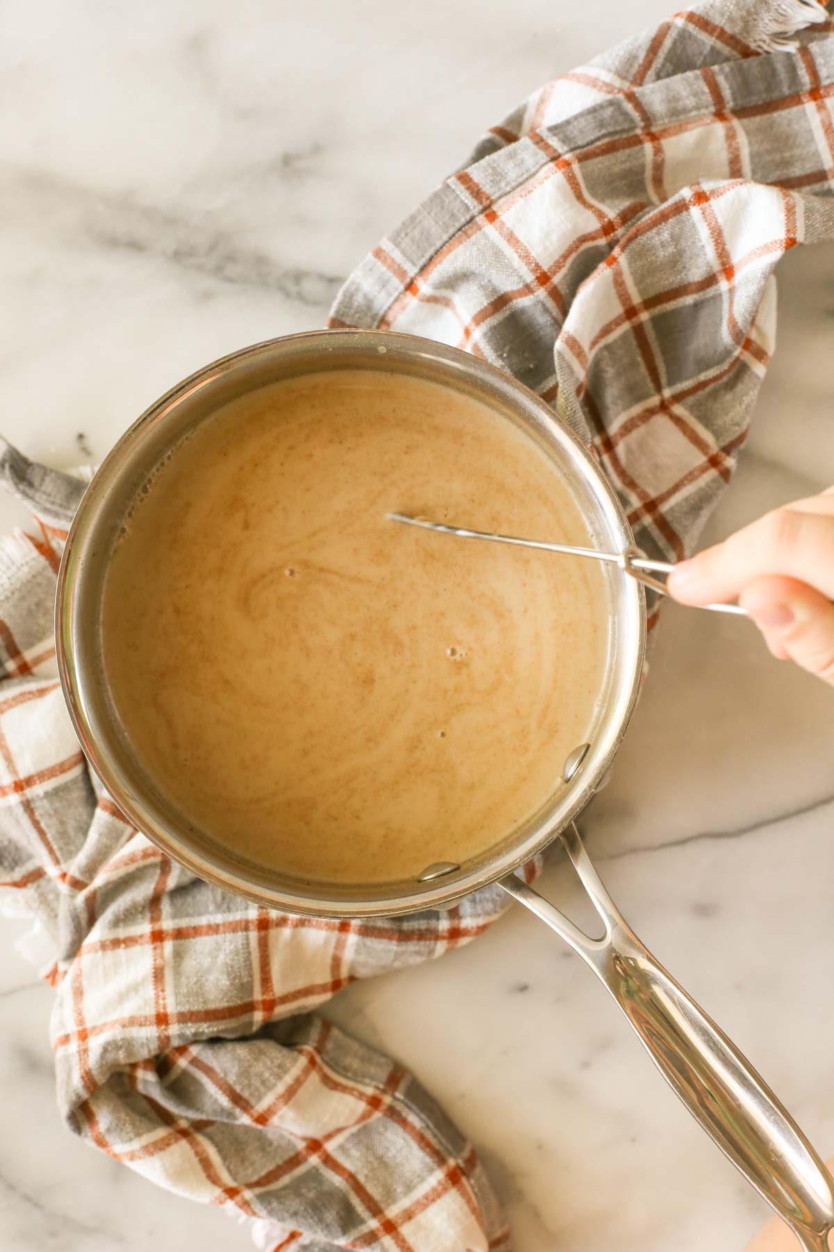 Overhead shot of a saucepan of Brown Sugar and Cinnamon Coffee Creamer with a hand holding on to a whisk in the saucepan, and the saucepan is sitting on a plaid cloth on a marble background.