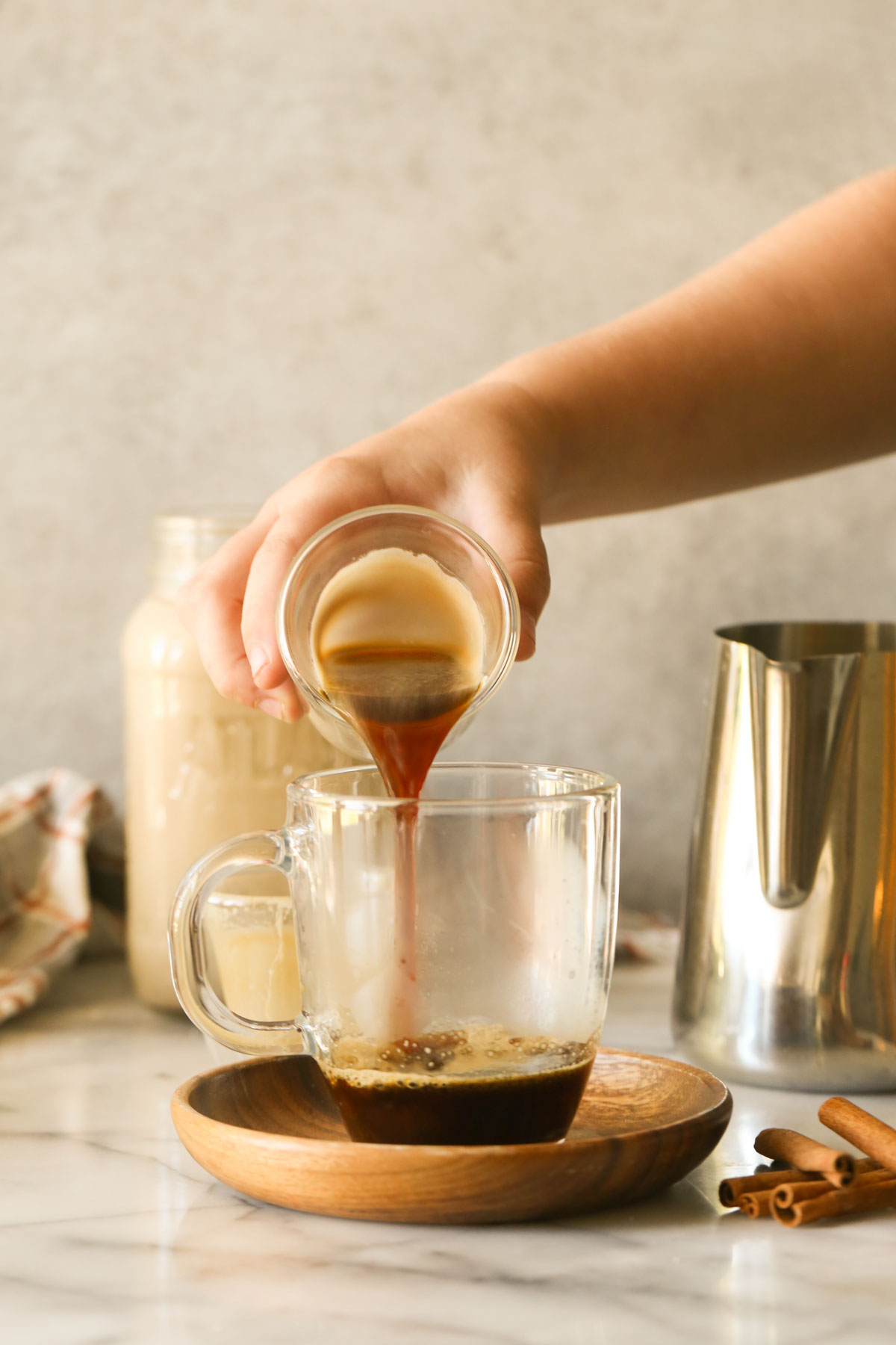 A hand pouring an espresso shot into a glass mug on a wood saucer with cinnamon sticks next to it, with a glass jar of Brown Sugar and Cinnamon Coffee Creamer and a stainless steel pitcher of steamed milk in the background.