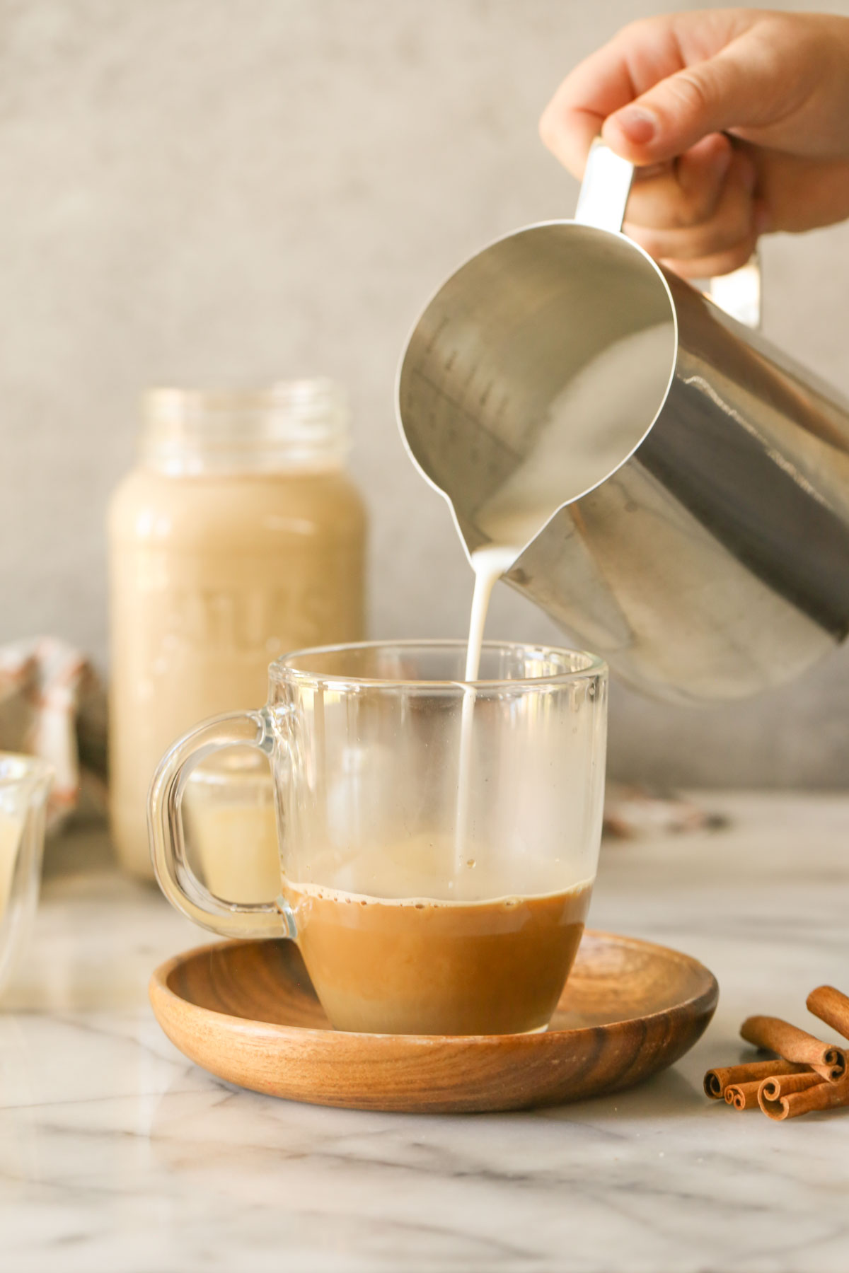 A hand pouring steamed milk from a stainless steel pitcher into a glass mug with espresso in it, sitting on a wood saucer with cinnamon sticks next to it, with a glass jar of Brown Sugar and Cinnamon Coffee Creamer in the background.