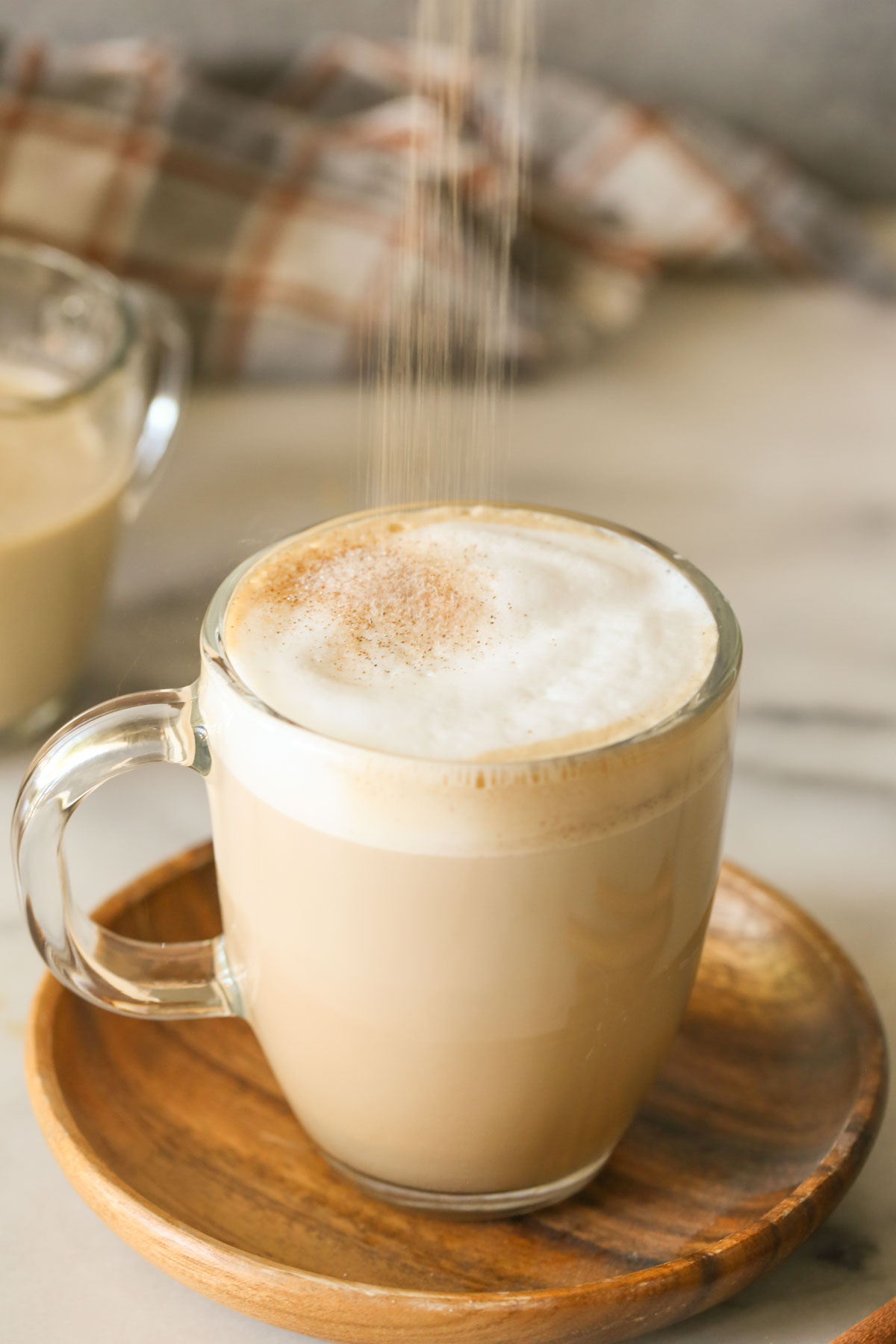 Cinnamon and sugar being dusted on top of a latte in a glass mug that has Brown Sugar and Cinnamon Coffee Creamer in it, that is sitting on a wood saucer.