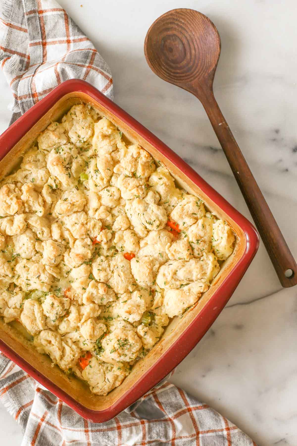 Overhead shot of a square baking dish of Chicken and Biscuits with a wooden serving spoon next to it, all on a marble background.