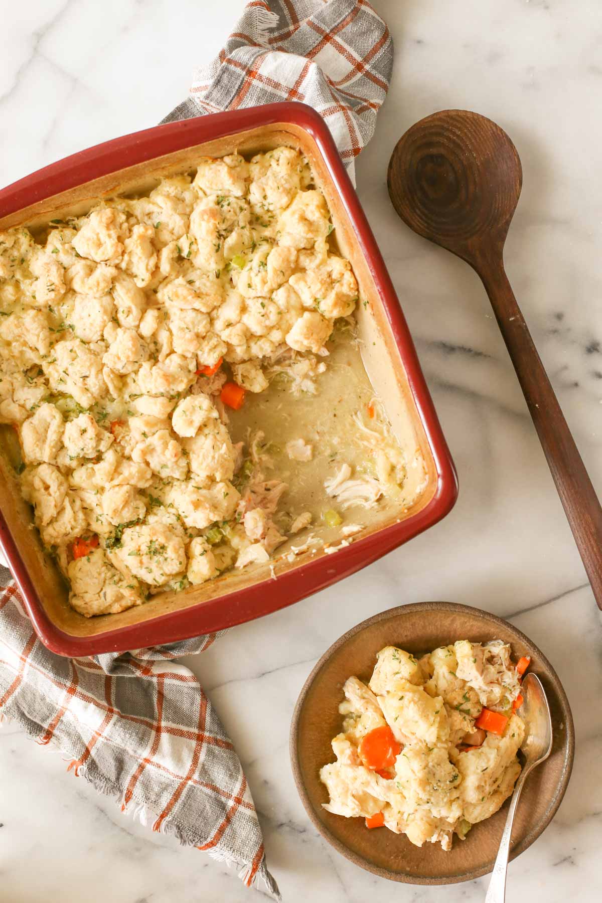 Overhead shot of a square baking dish of Chicken and Biscuits with some missing that has been served in a bowl next to the baking dish, with a wooden serving spoon next to the baking dish as well, all on a marble background.