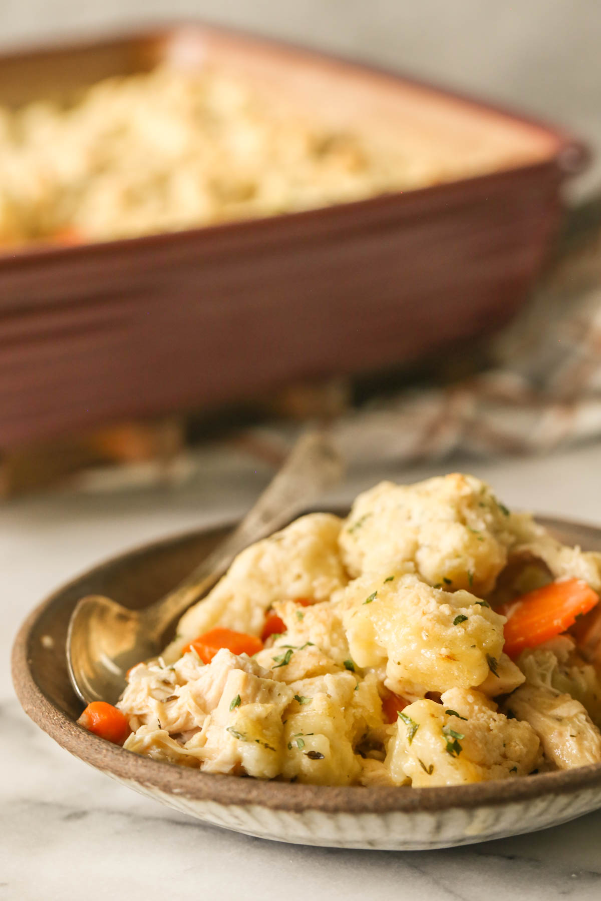 Close up shot of a serving of Chicken and Biscuits in a bowl with a spoon in it, and the baking dish of Chicken and Biscuits in the background.