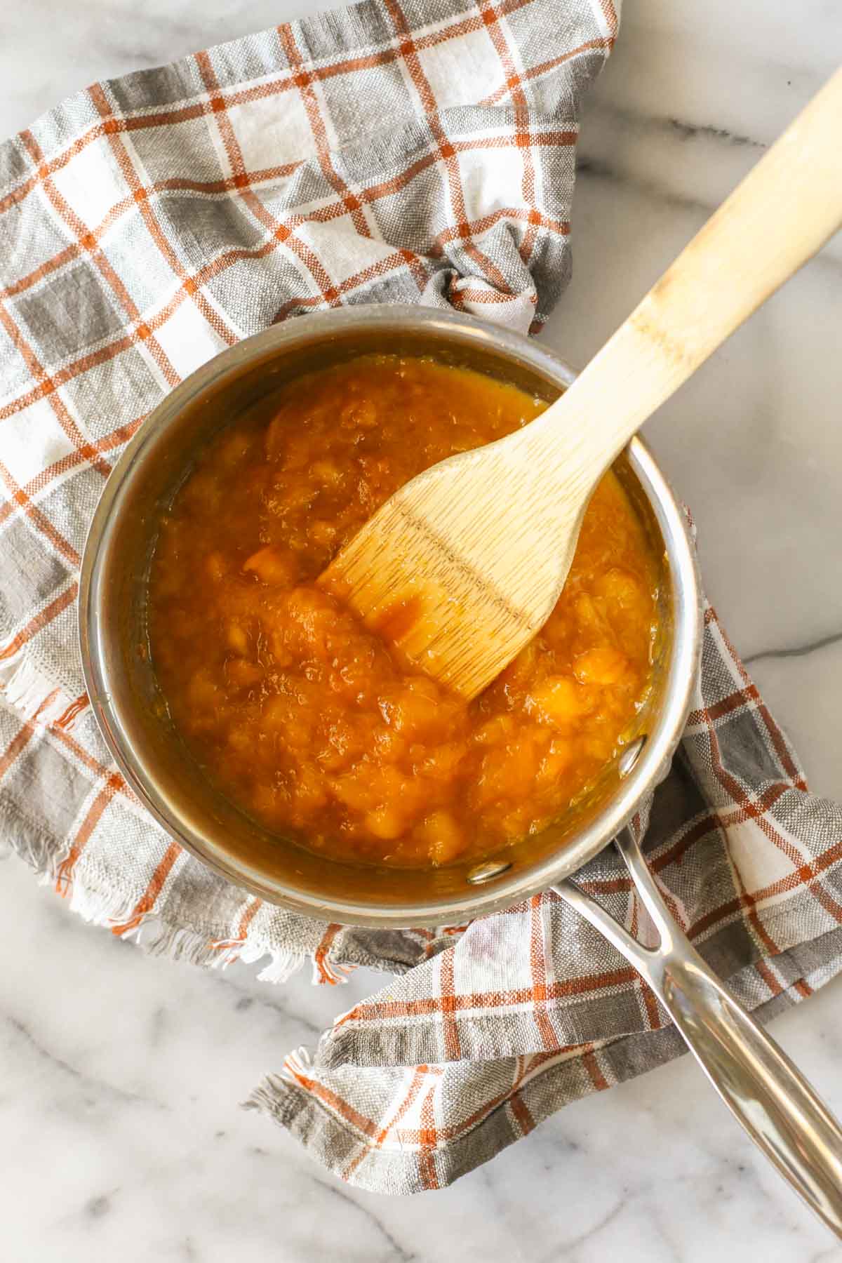Overhead shot of a saucepan of the peach filling for Peach Crumb Bars, sitting on a plaid dish towel with a wooden spoon in the filling.  