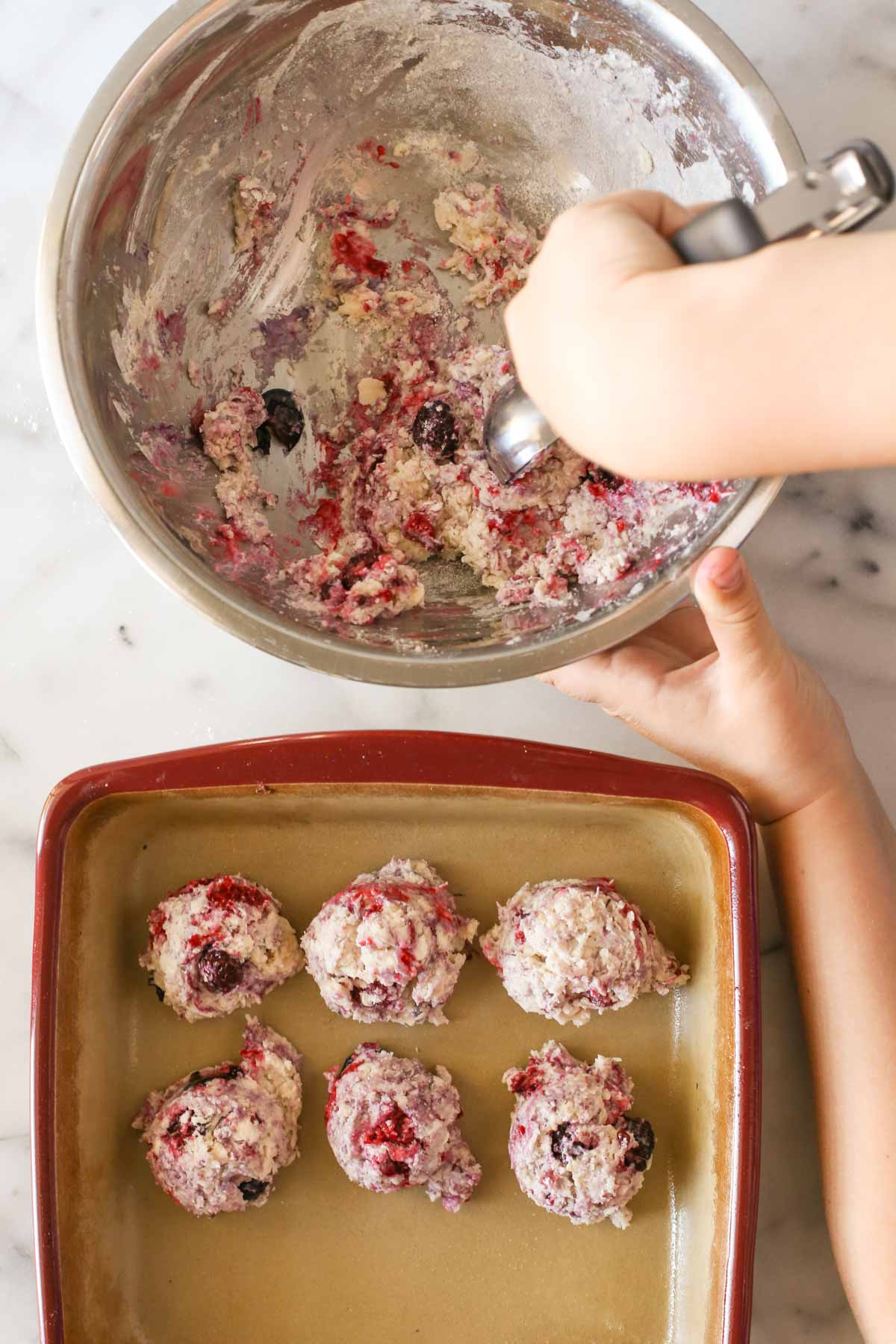 Overhead shot of a hand scooping out the Glazed Berry Drop Biscuit dough from a mixing bowl that is sitting next to a baking pan that has six Glazed Berry Drop Biscuit dough mounds in it.