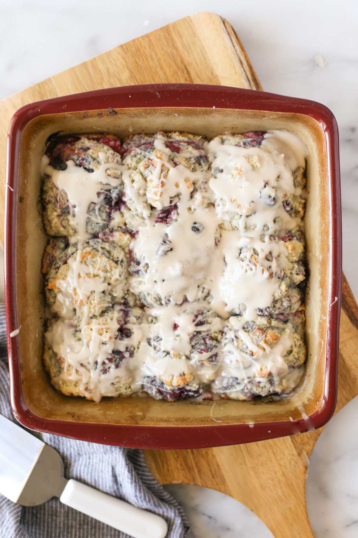 Overhead shot of a baking pan of Glazed Berry Drop Biscuits with the glaze on top, sitting on a wood cutting board, with a serving spatula next to it.