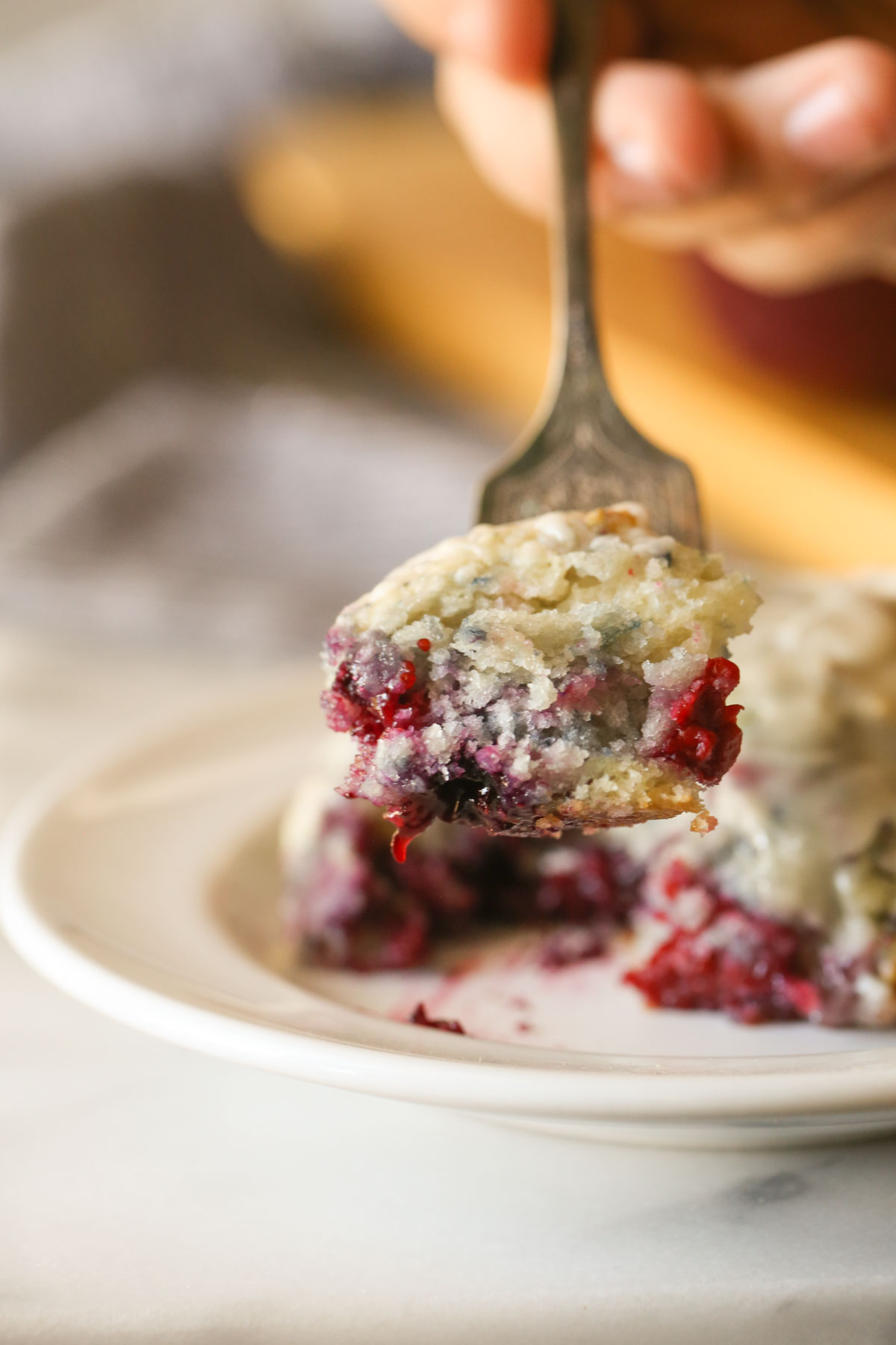 Close up shot of a fork with a bite of a Glazed Berry Drop Biscuit on it, with a plate of Glazed Berry Drop Biscuits in the background.